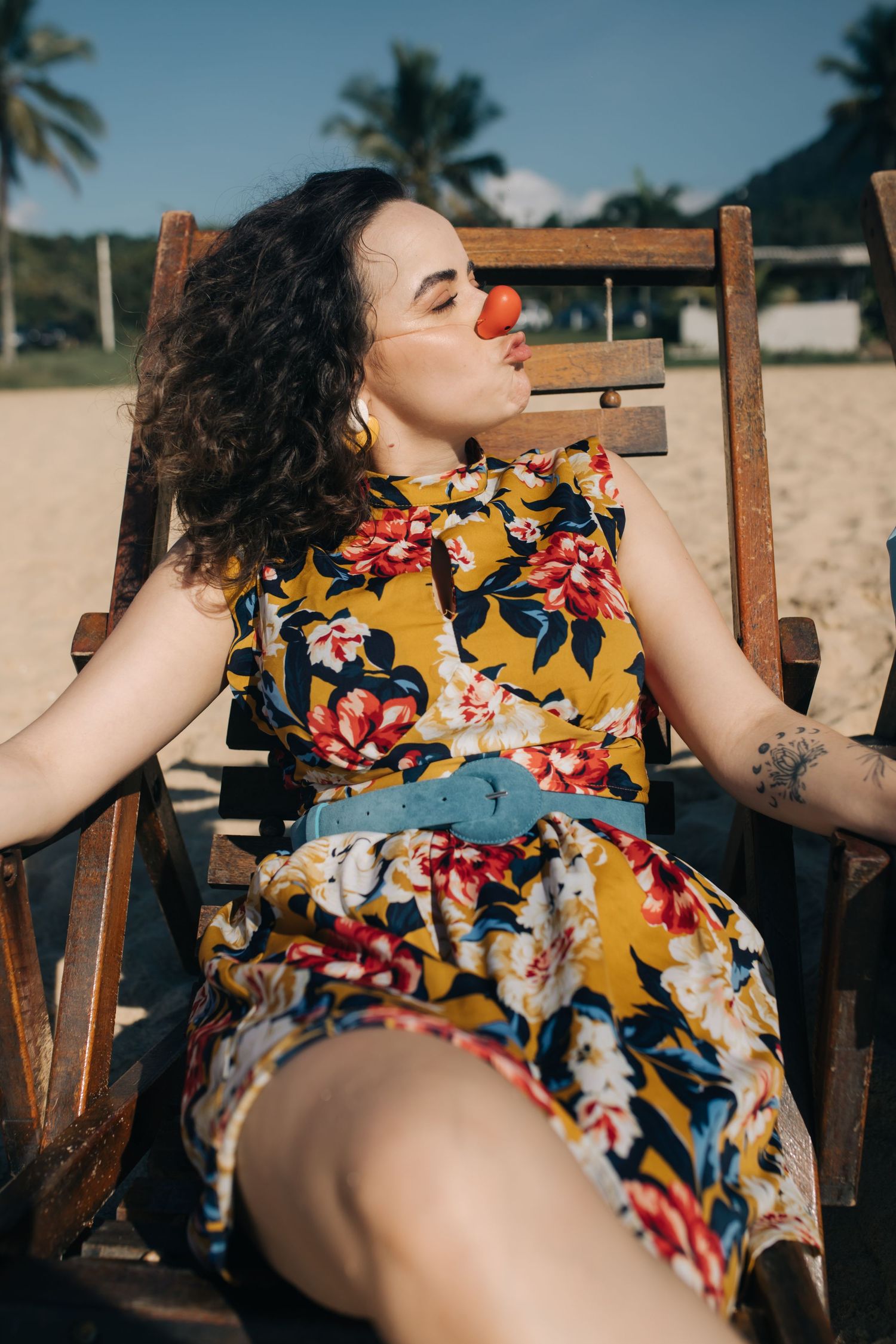 Person in floral dress relaxing in wooden beach chair with palm trees in background.