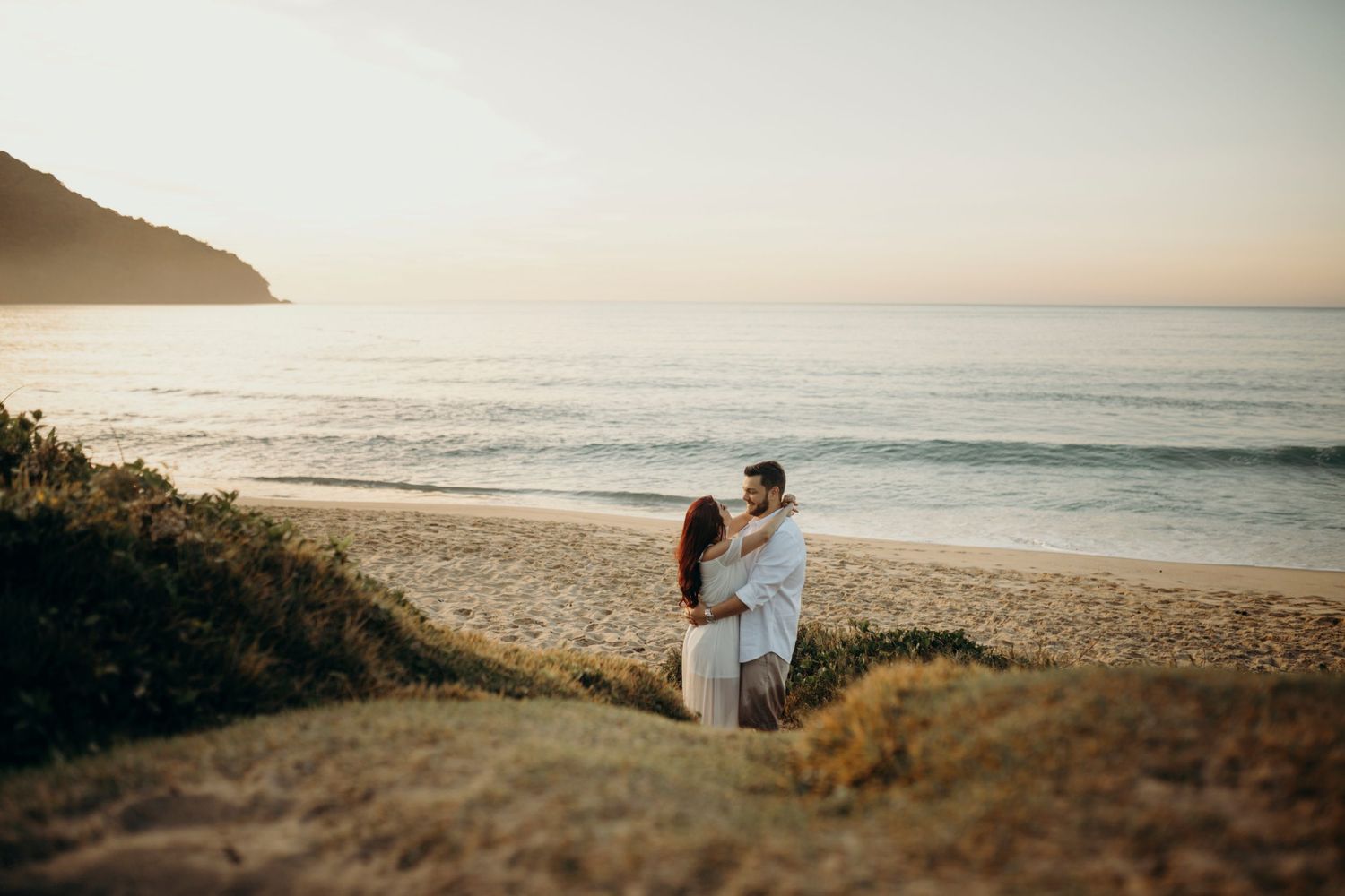 A couple embraces while standing on a grassy overlook above a scenic beach.