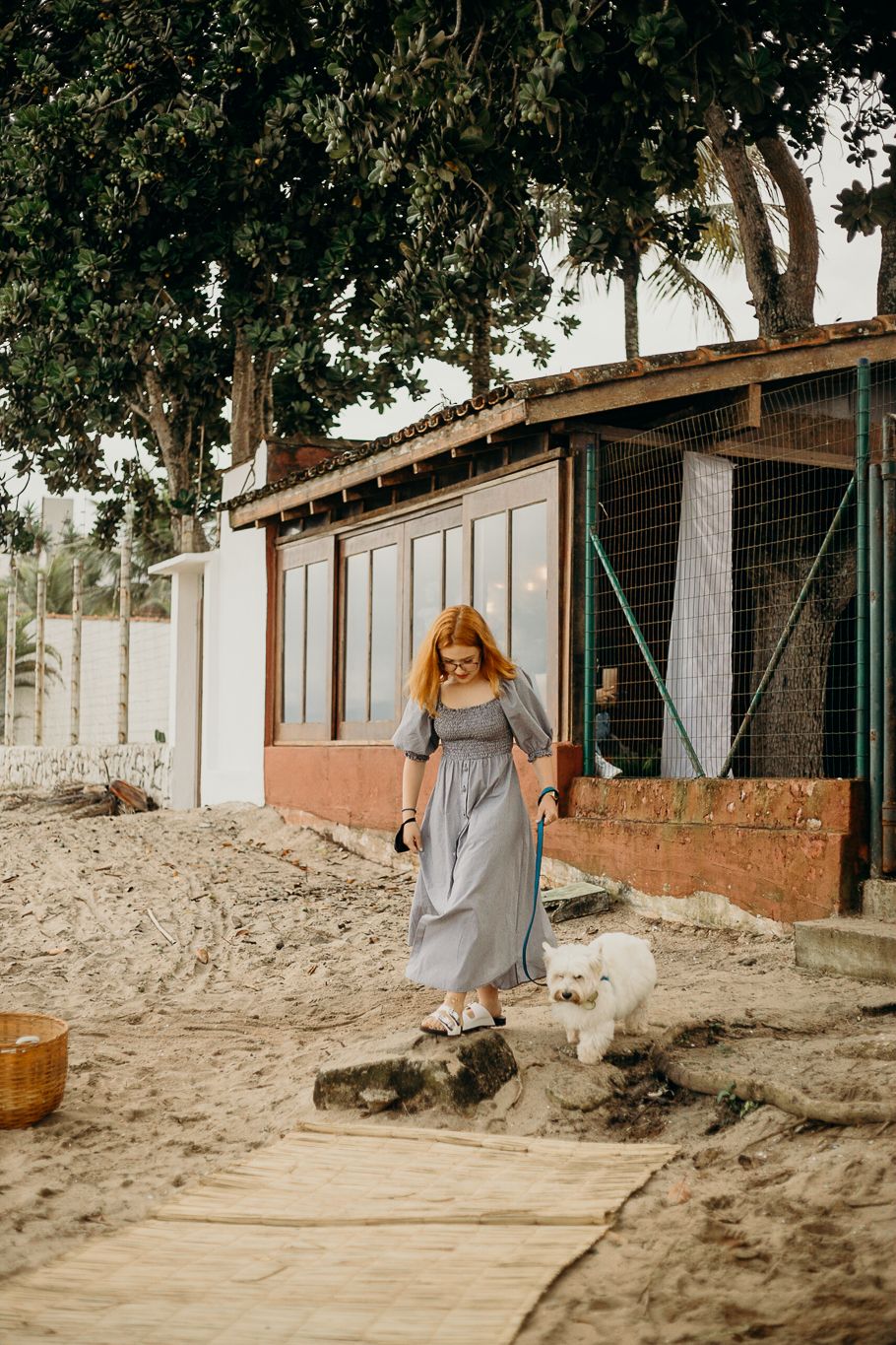 Person in a flowing gray dress walks along a sandy path with a small white dog.