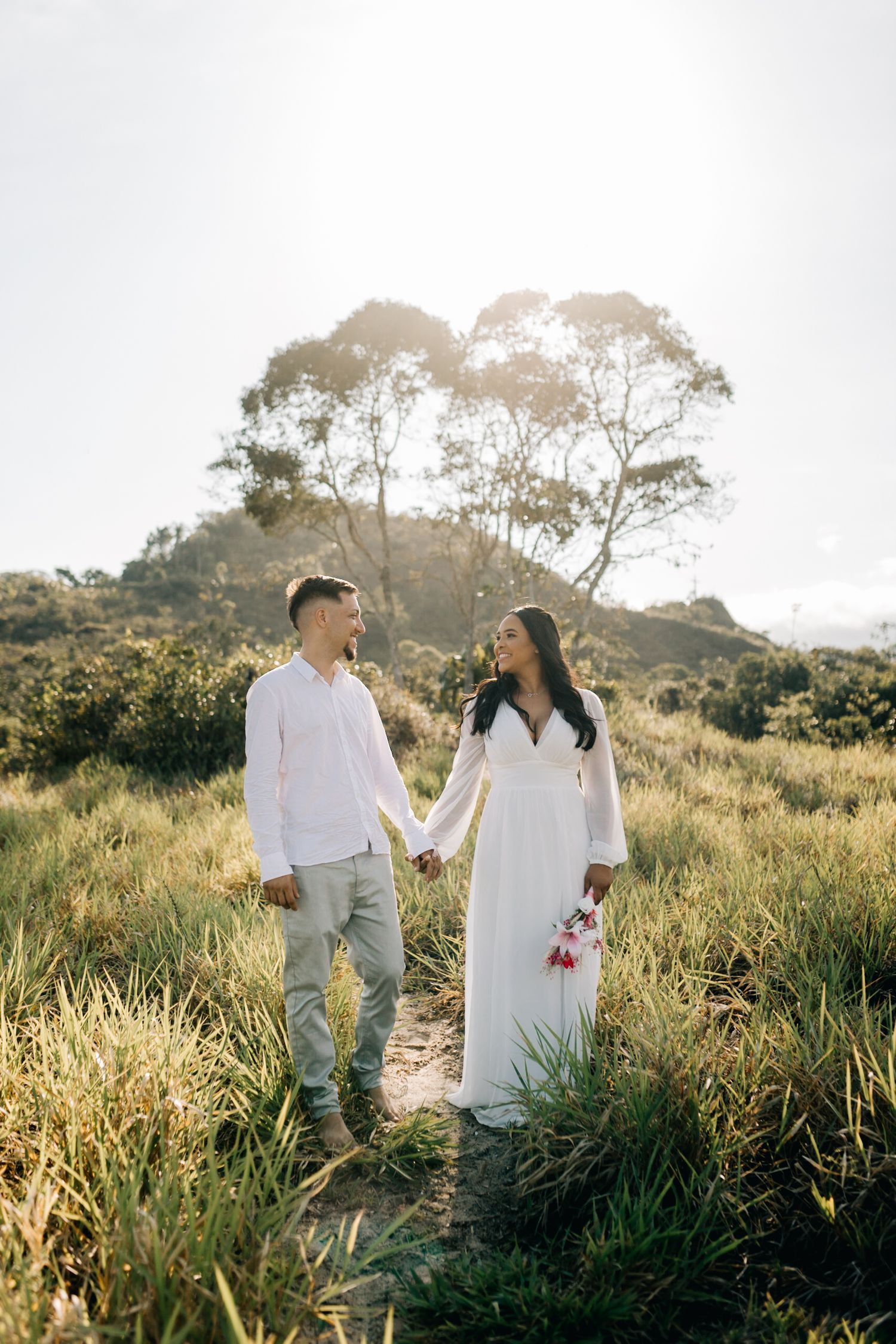 Couple in white outfits walking through tall grass on a hillside during golden hour.