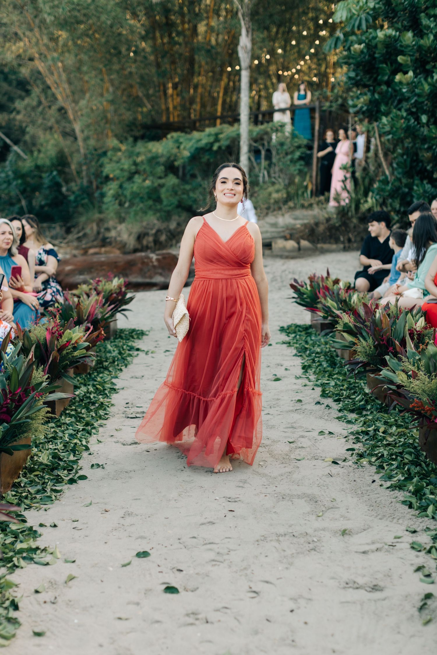 Bridesmaid in red chiffon dress walks garden path at outdoor wedding ceremony.