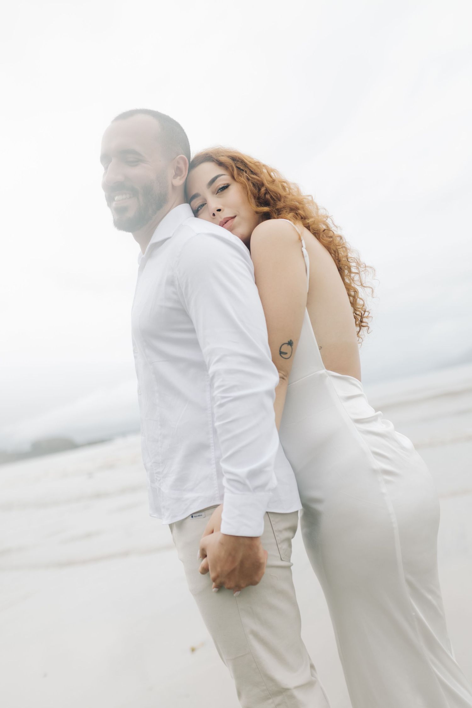 A couple in white clothing embraces intimately during a cloudy beach photoshoot.