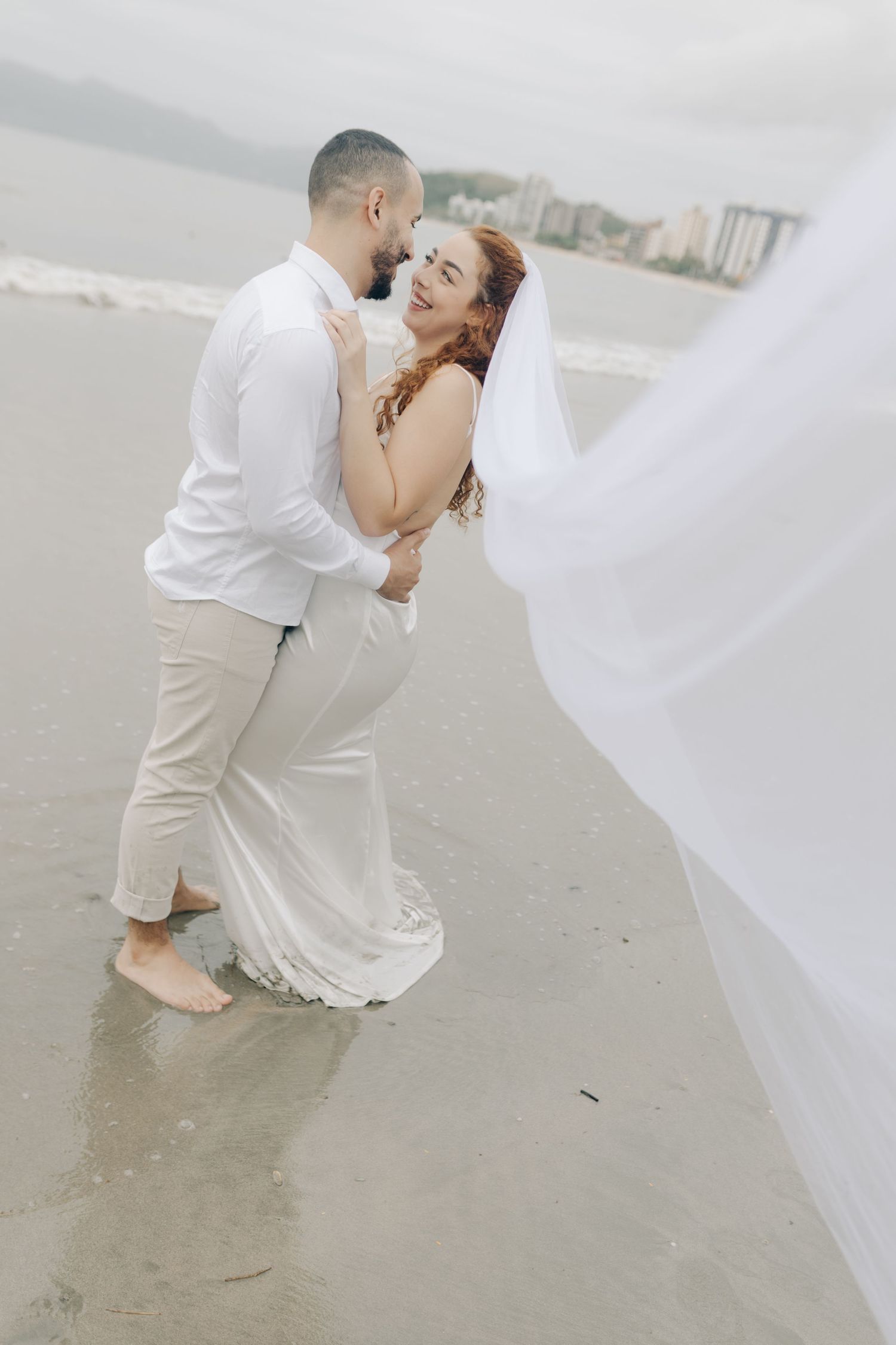 Wedding couple embracing on misty beach with flowing veil.