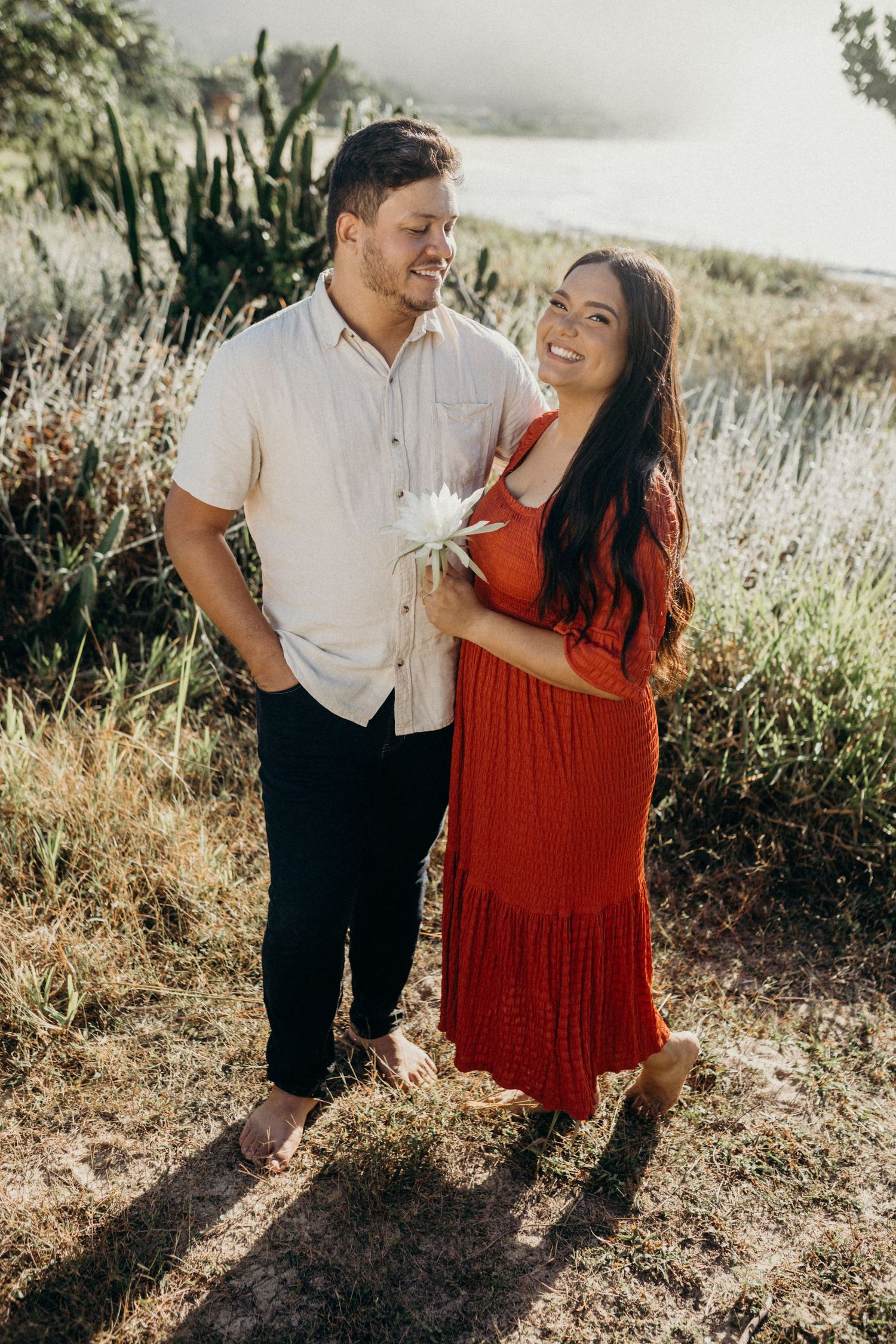 Happy couple in casual attire poses on hiking trail with mountain and water views.