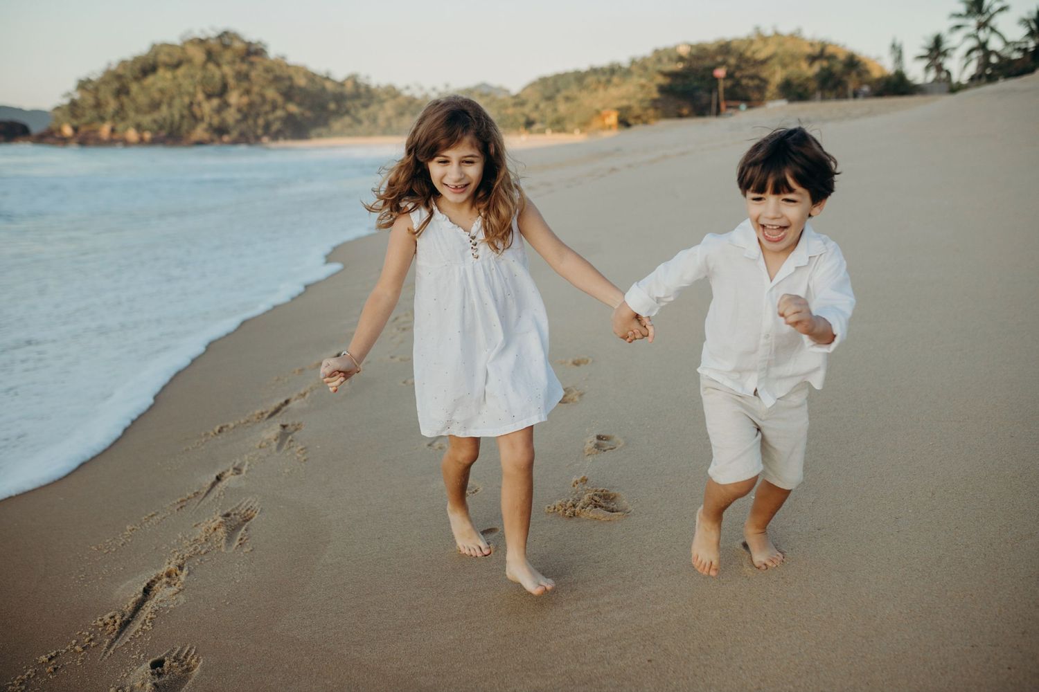 Two people in white clothing run playfully along a scenic beach at sunset, leaving footprints in the sand.