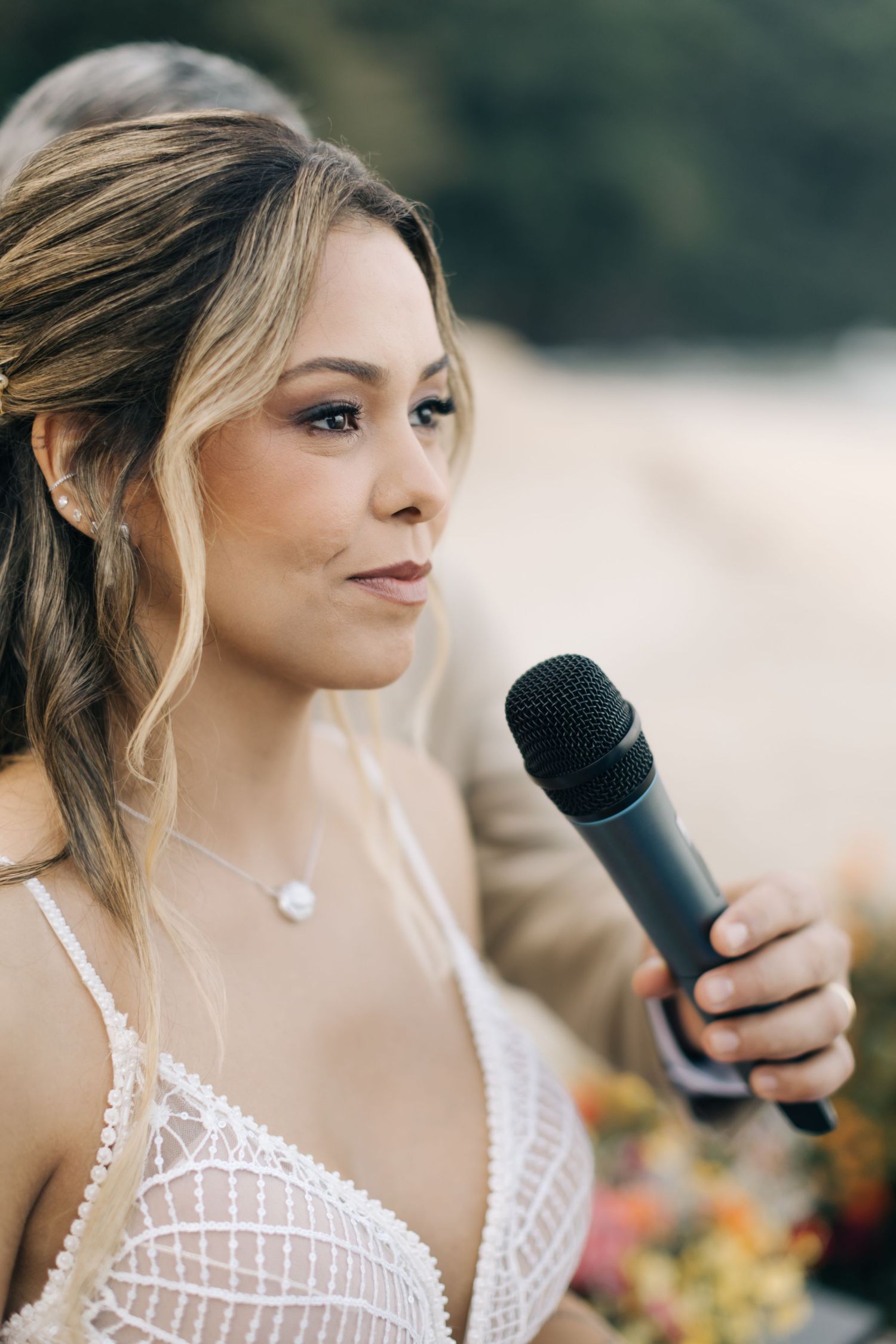 Bride in lace dress speaks into microphone during beachside wedding ceremony.