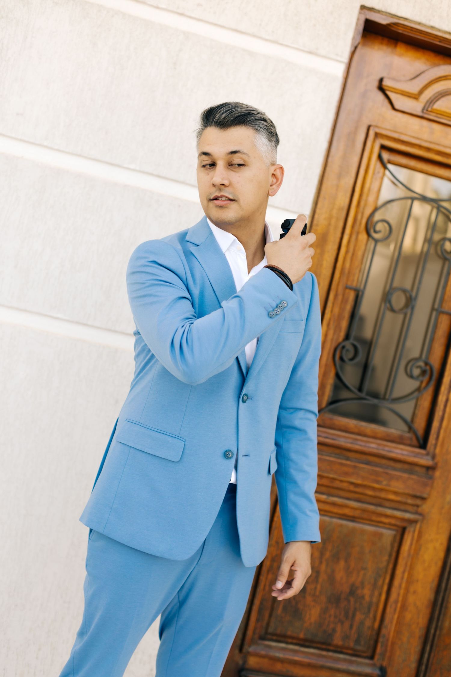 Person in powder blue suit adjusting lapel while posing against white wall and wooden door.