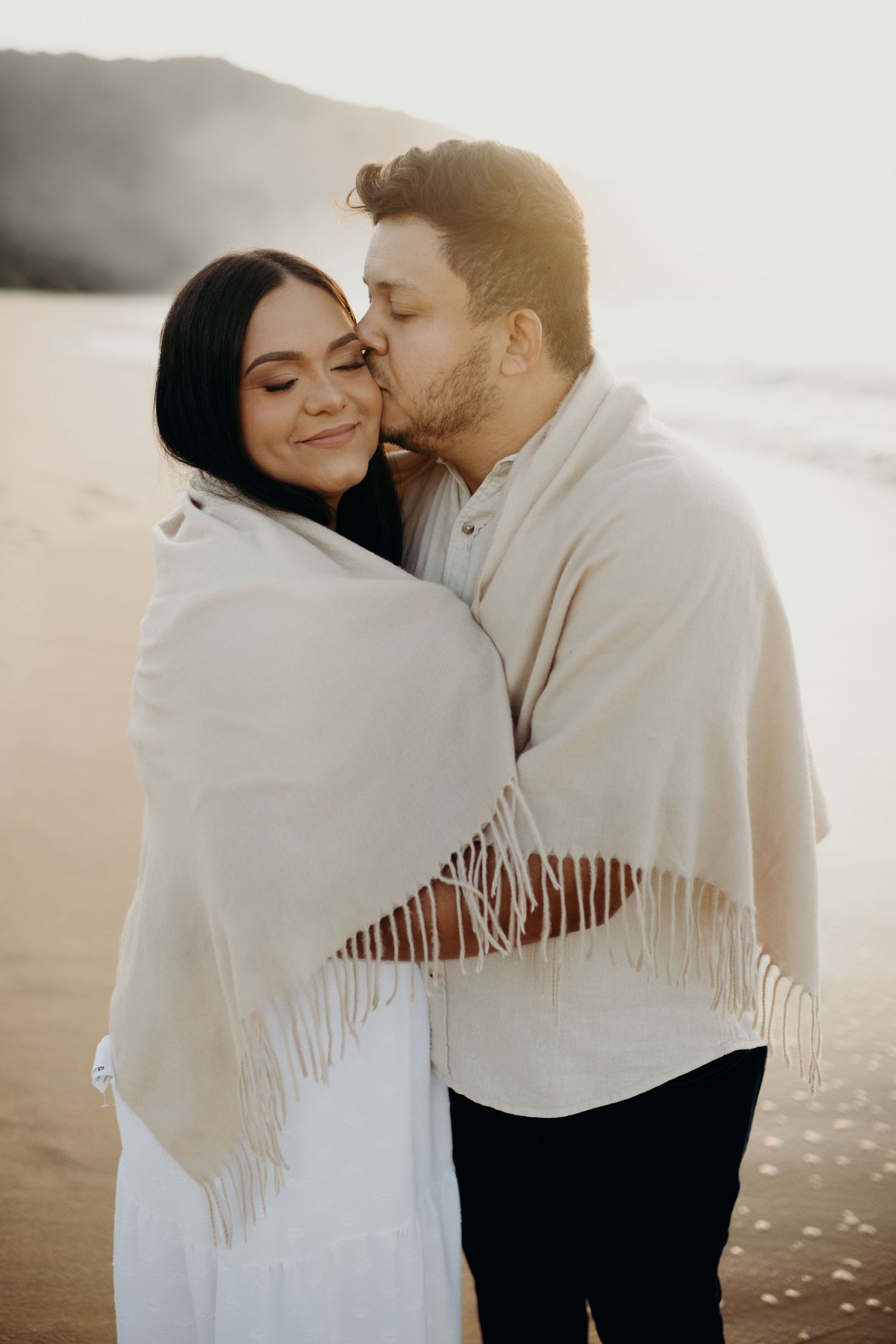 Close-up of couple wrapped in white shawl sharing tender moments on the beach.