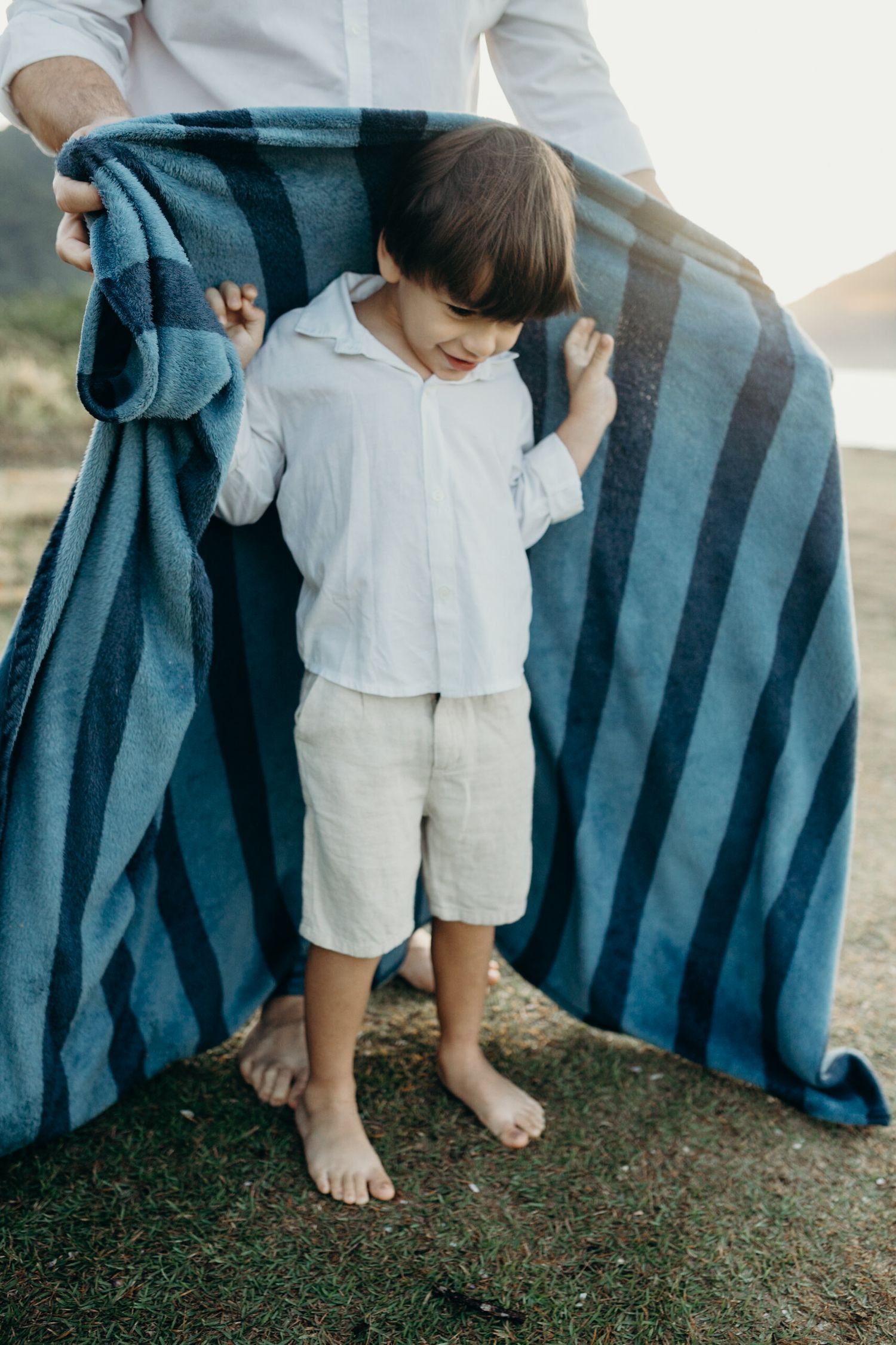 People playing peek-a-boo with a blue striped beach towel on grassy ground.
