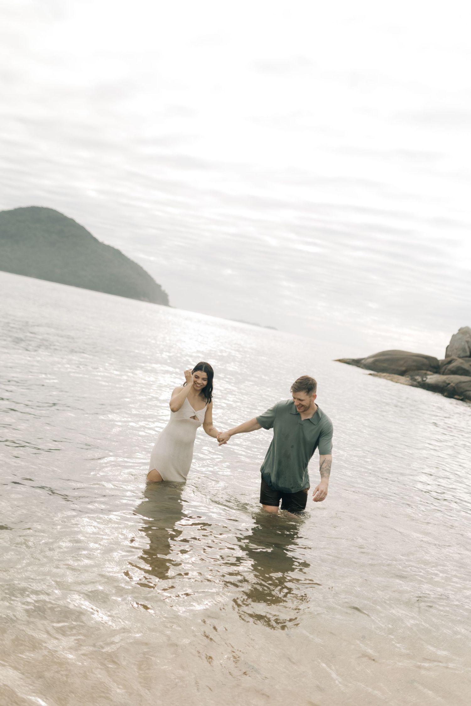 A couple holds hands while wading through ocean water near a rocky coastline.