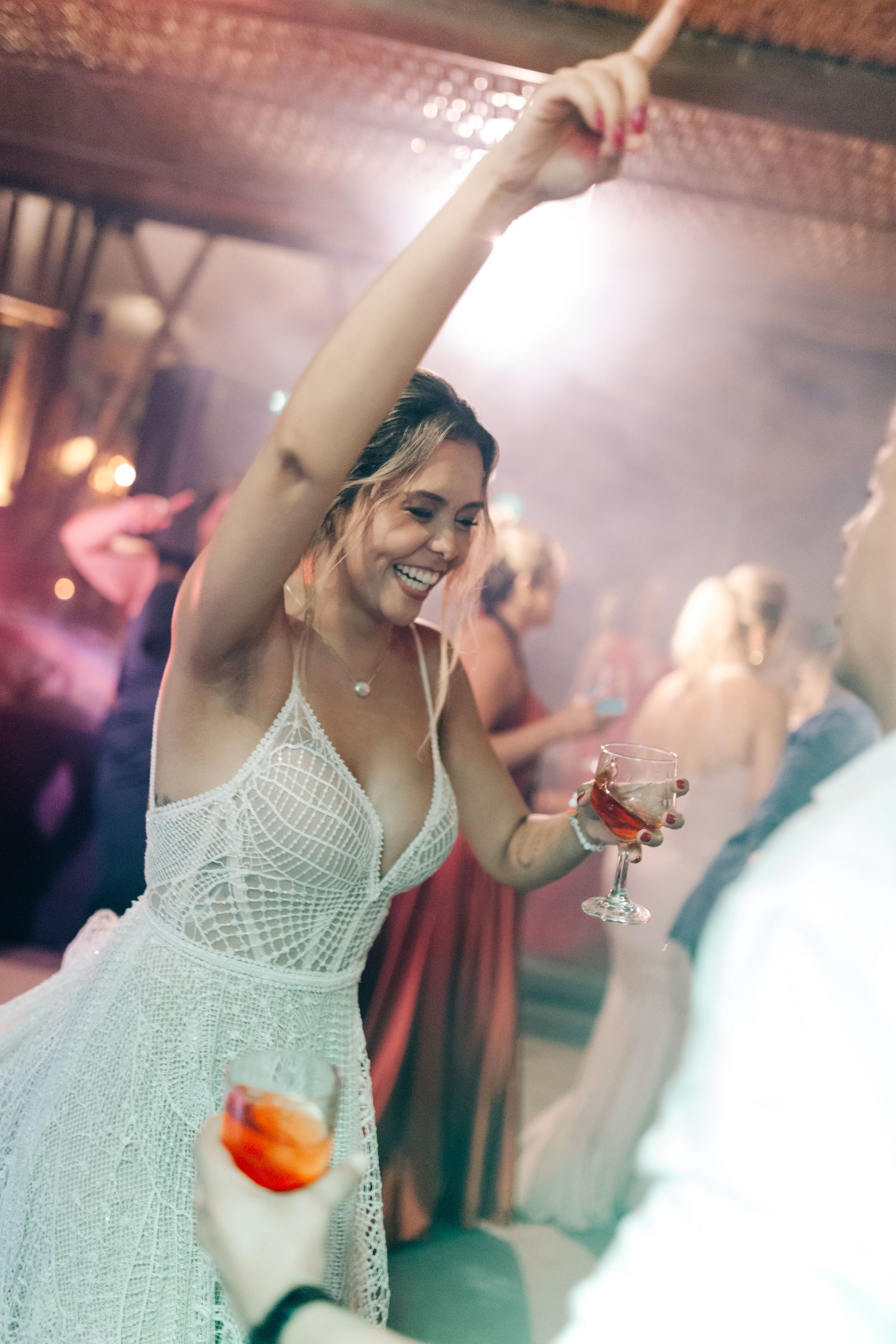 Bride in lace dress dancing joyfully at reception while holding a cocktail.