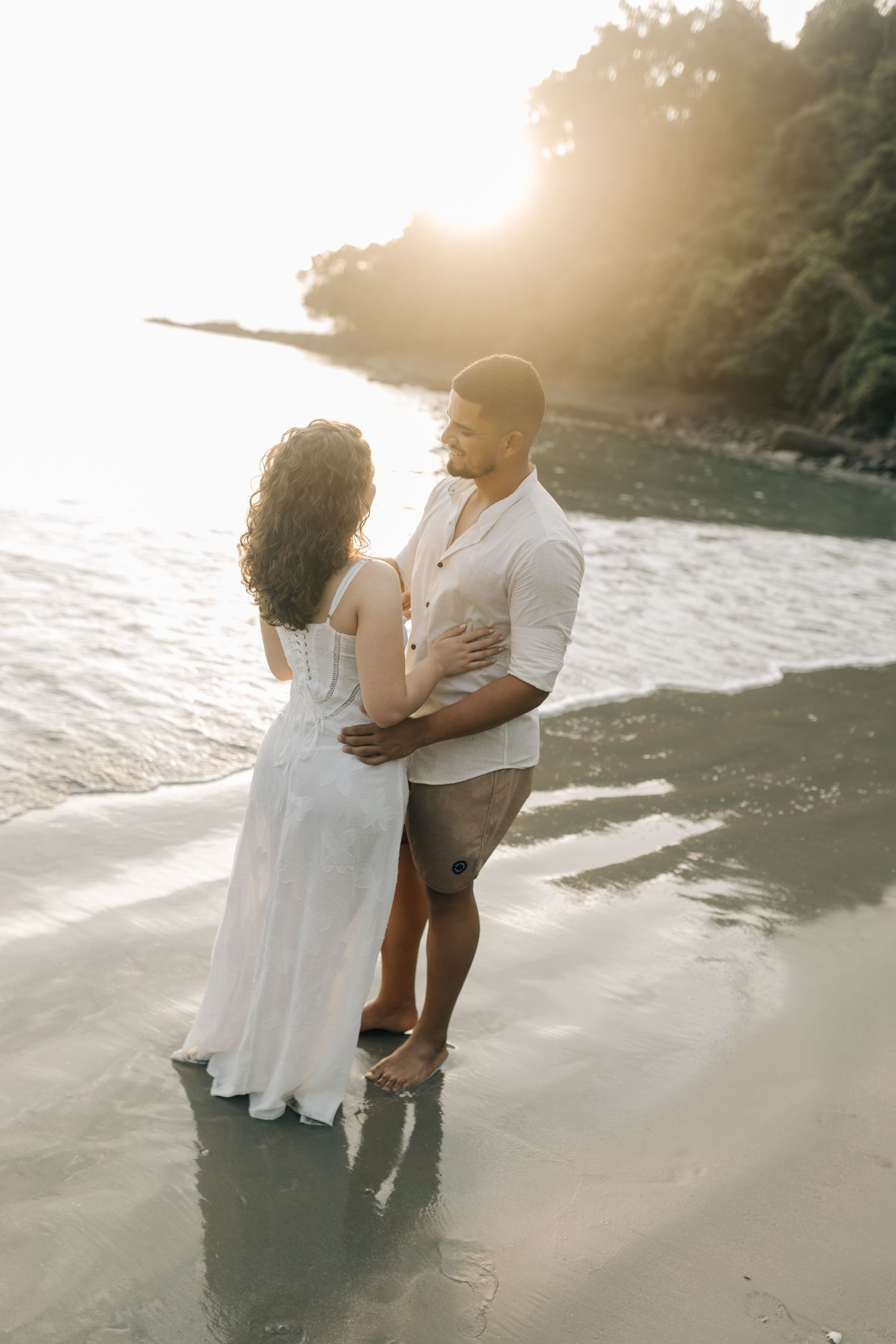 A romantic couple embraces on a sun-drenched beach during a sunset photoshoot.