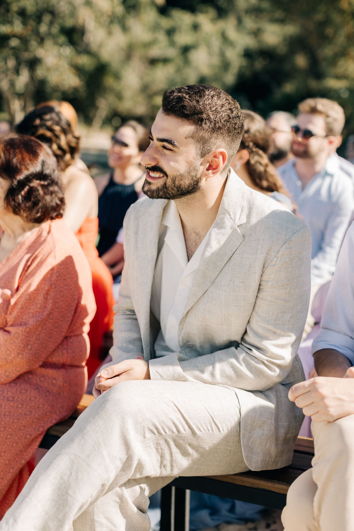 Wedding guests laugh and enjoy themselves while seated at an outdoor ceremony in a garden setting.