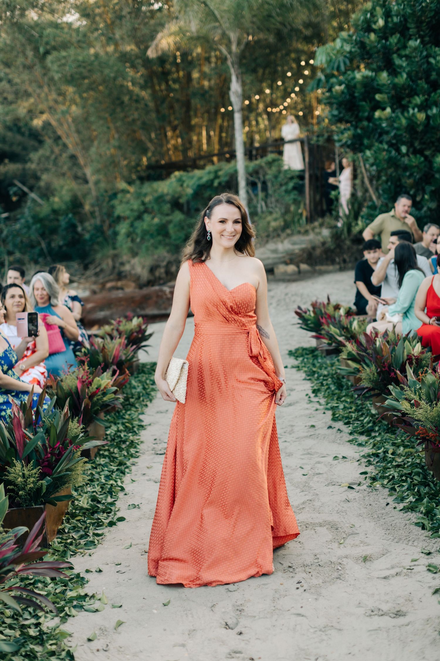 Person in a coral-colored dress walking down a sandy aisle lined with tropical plants.