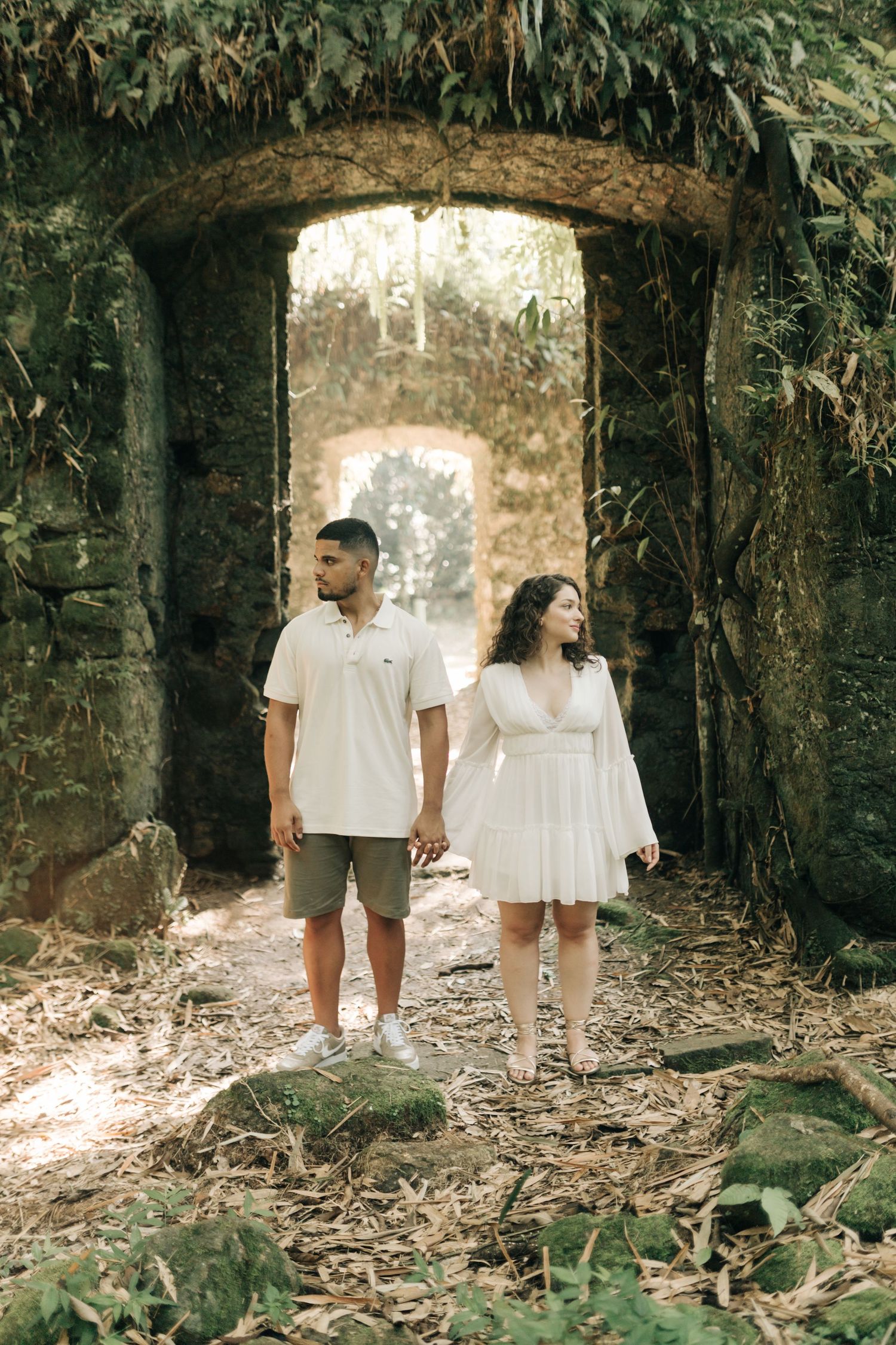 Couple takes romantic stroll through stone archway in forest wearing coordinated white ensembles.