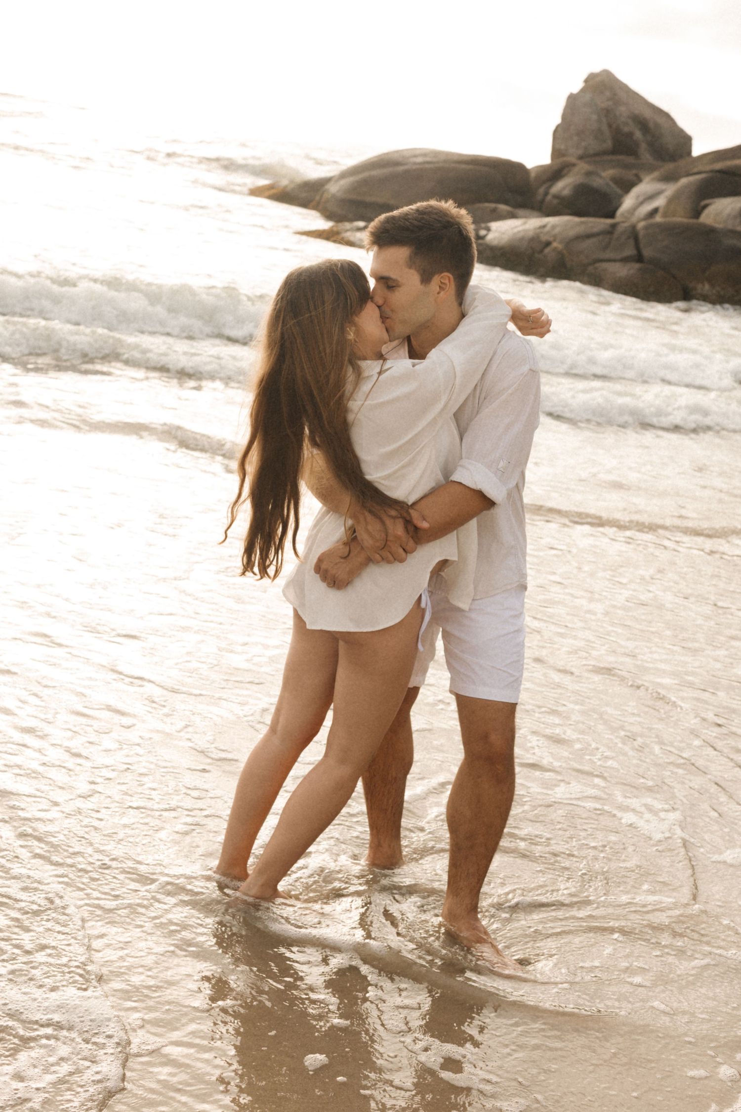 A romantic couple in white outfits embraces while standing in shallow ocean water at sunset near rocky coastline.