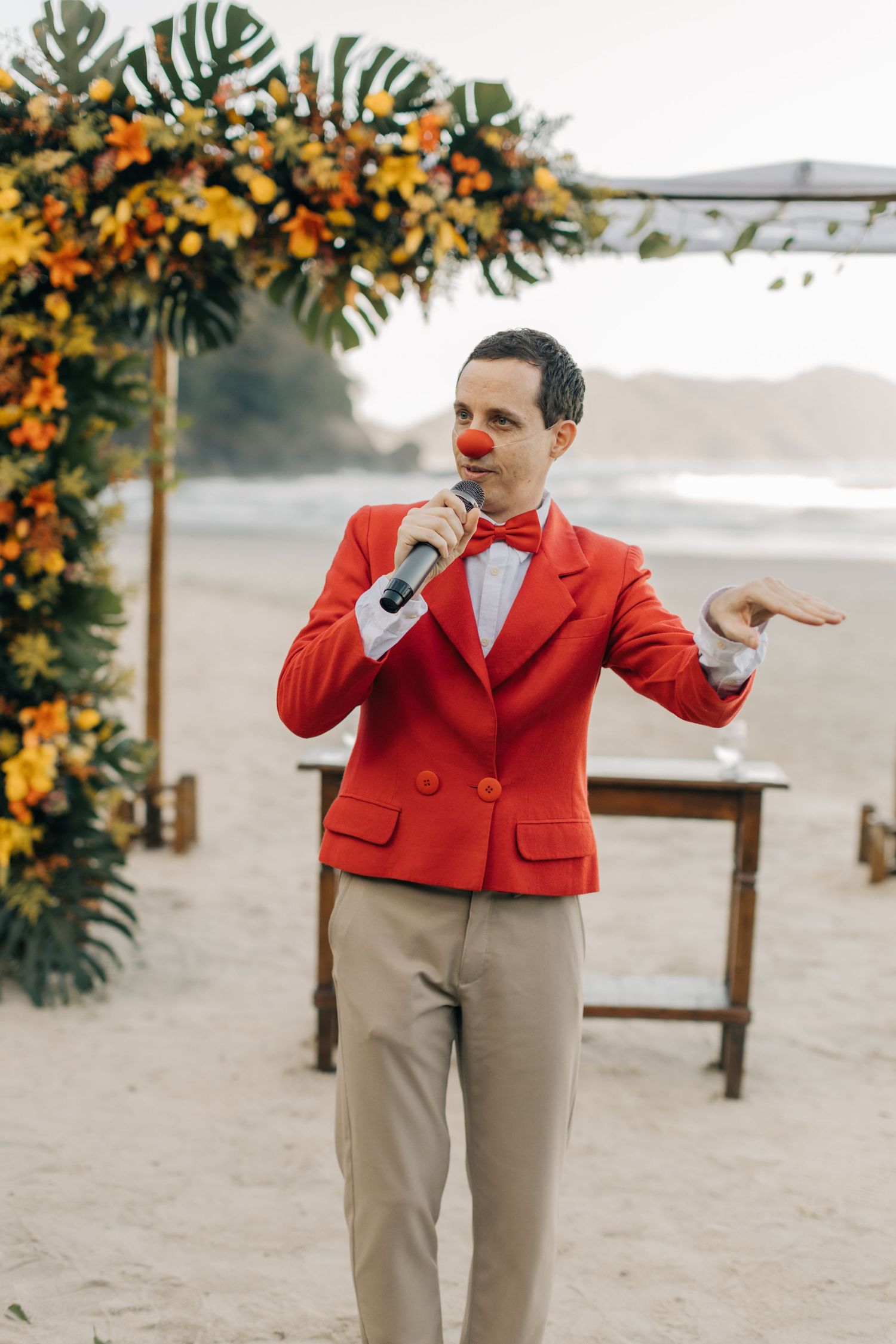 A performer in a bright red blazer entertains at a beach wedding under a floral arch.