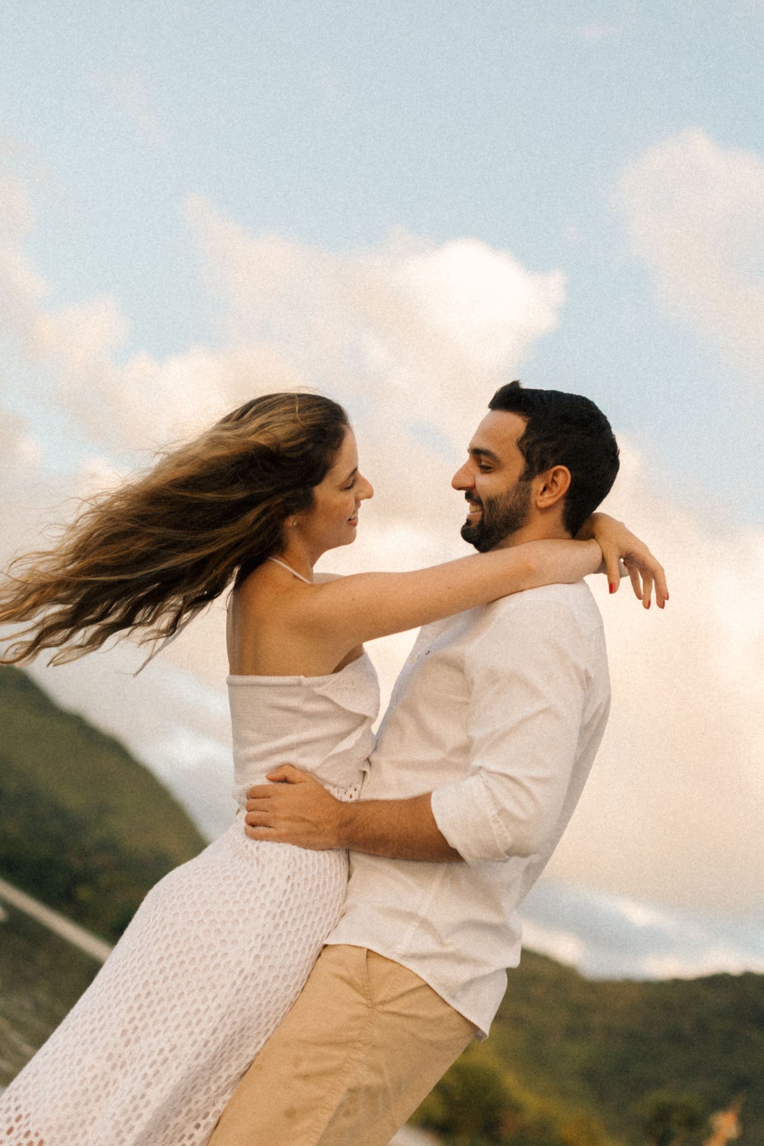 Playful couple in white outfits sharing joyful moment on beach with mountains in background.
