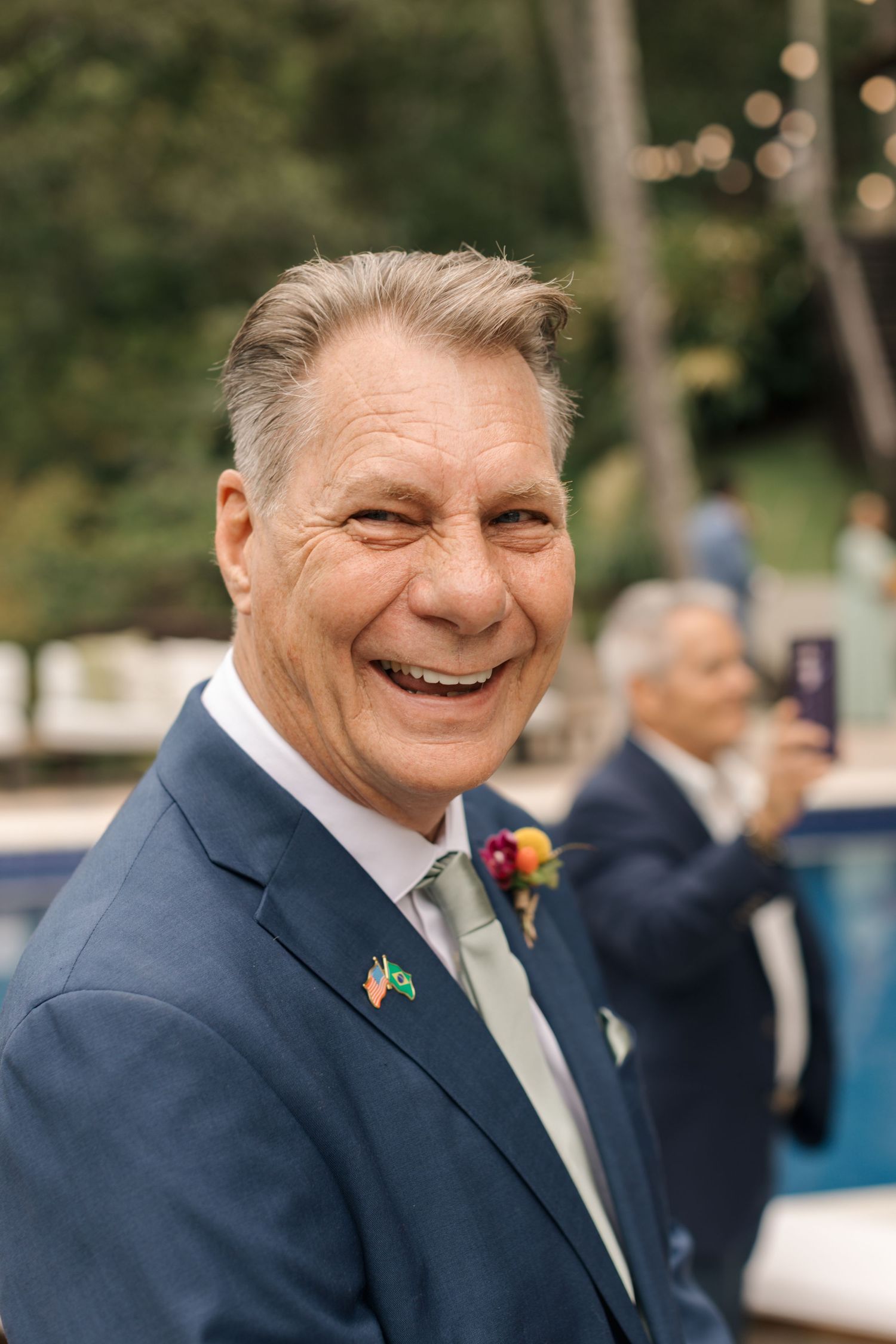 Wedding guest in a navy suit smiling joyfully at an outdoor celebration.