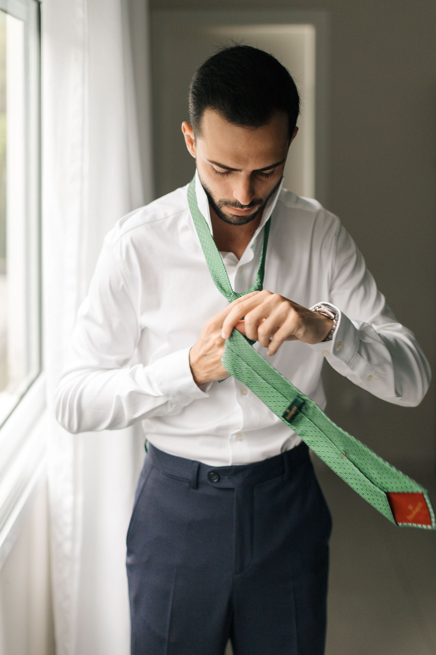 Person adjusting a green necktie while wearing crisp white shirt and navy trousers.