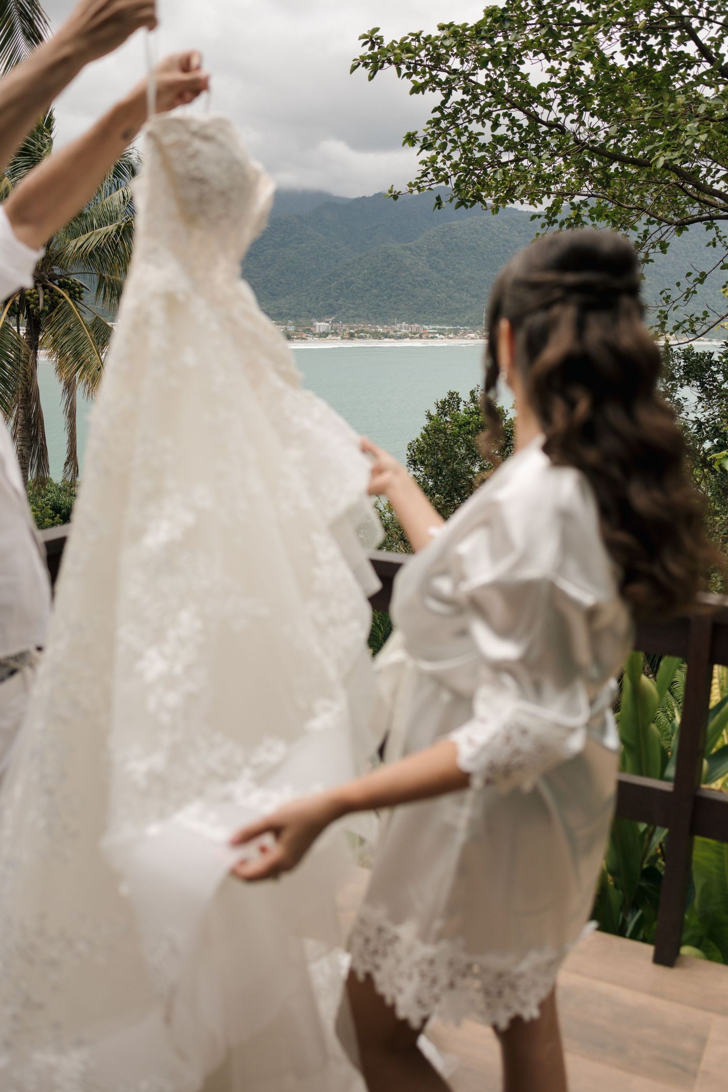 A white lace wedding dress hangs while being prepared with a scenic coastal mountain view in the background.