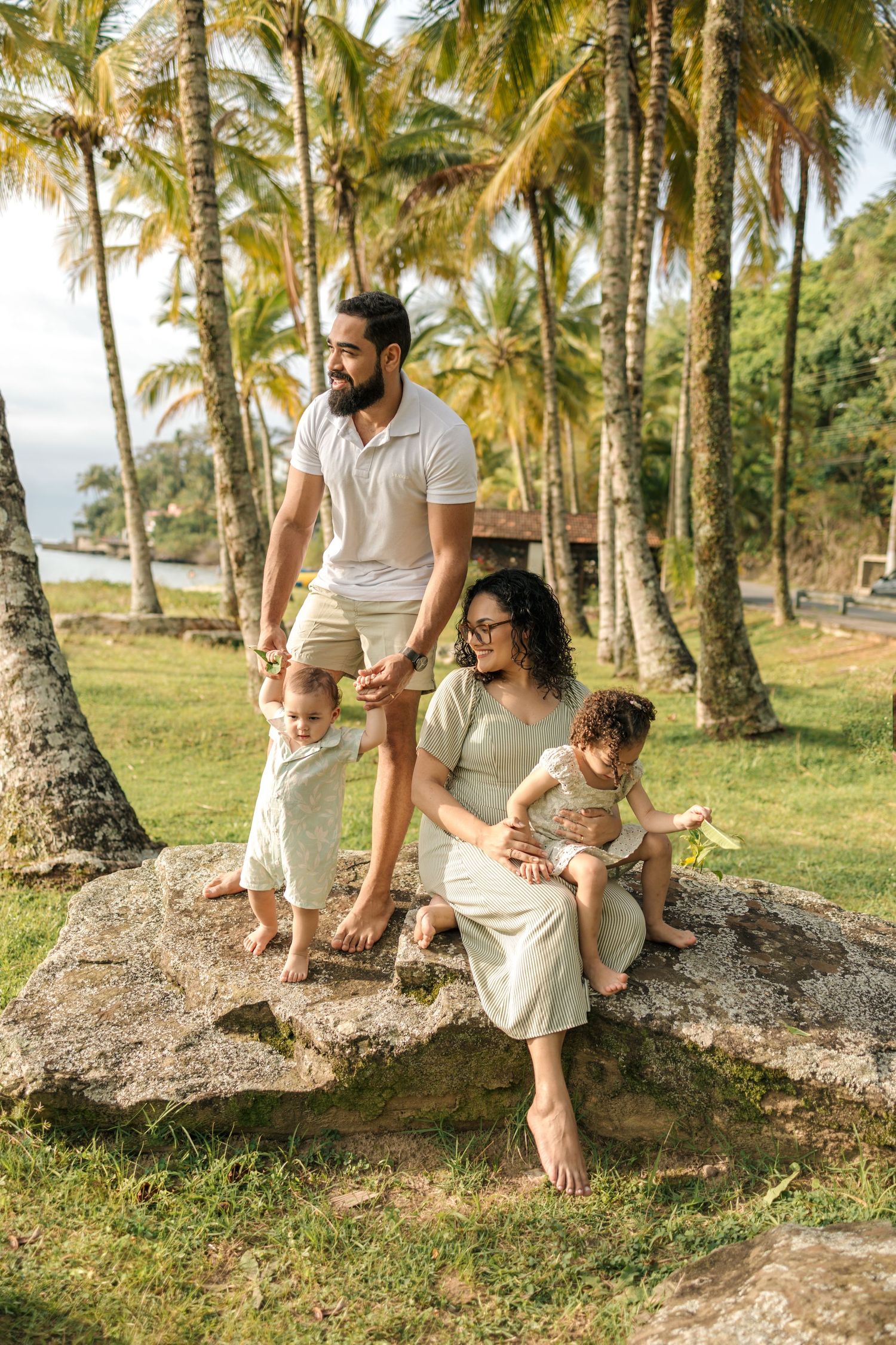 A family enjoys a relaxing moment together sitting on palm tree roots in a tropical setting wearing light colored clothing.