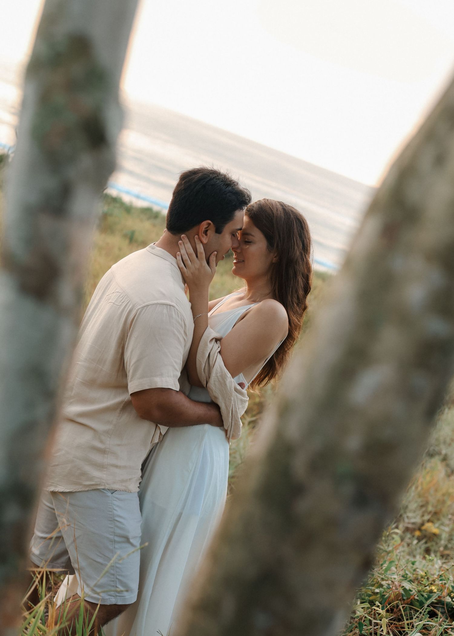 Romantic couple in white clothing embraces between trees at golden hour.
