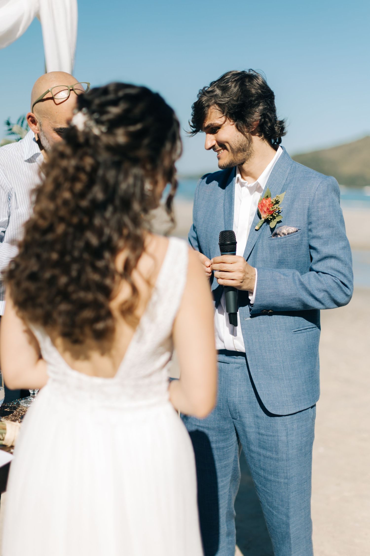 Groom in light blue suit speaks into microphone during beach wedding ceremony.