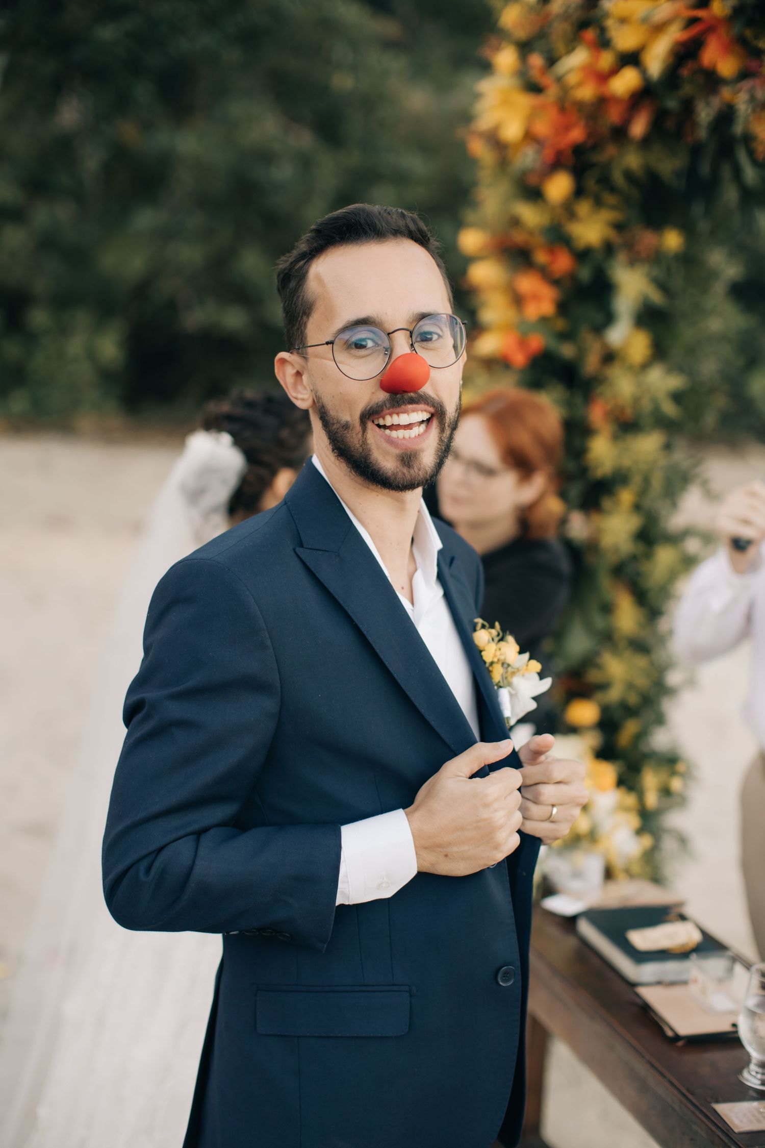 Groom wearing red nose and navy suit laughs during beach wedding ceremony.