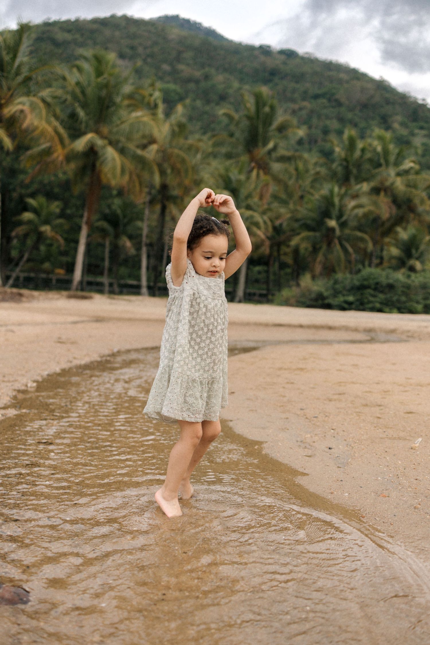 A small child in a white dress plays and dances at the edge of the water on a tropical beach with palm trees.