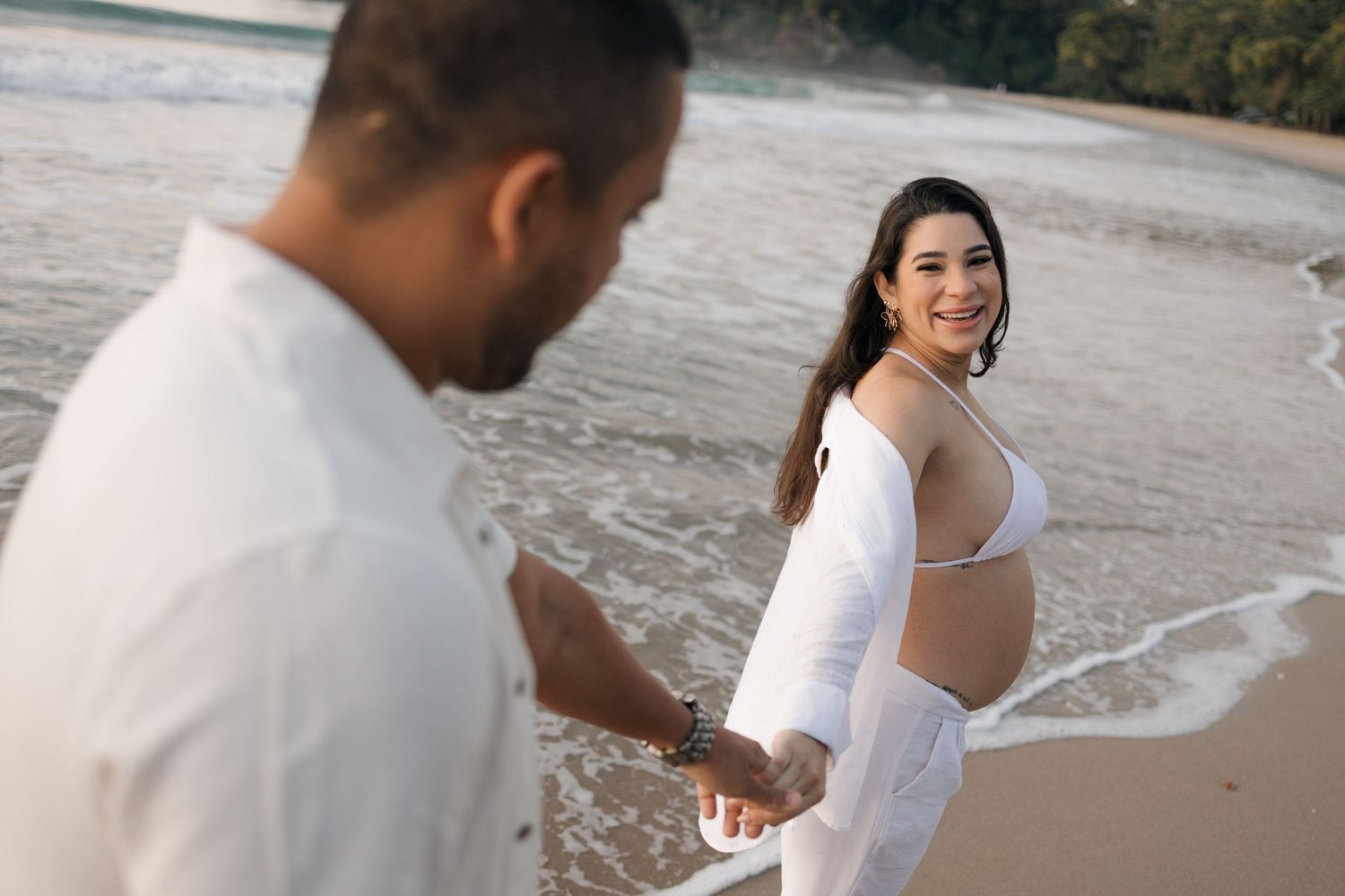 Couple holding hands on beach with waves lapping at their feet during sunset photoshoot.