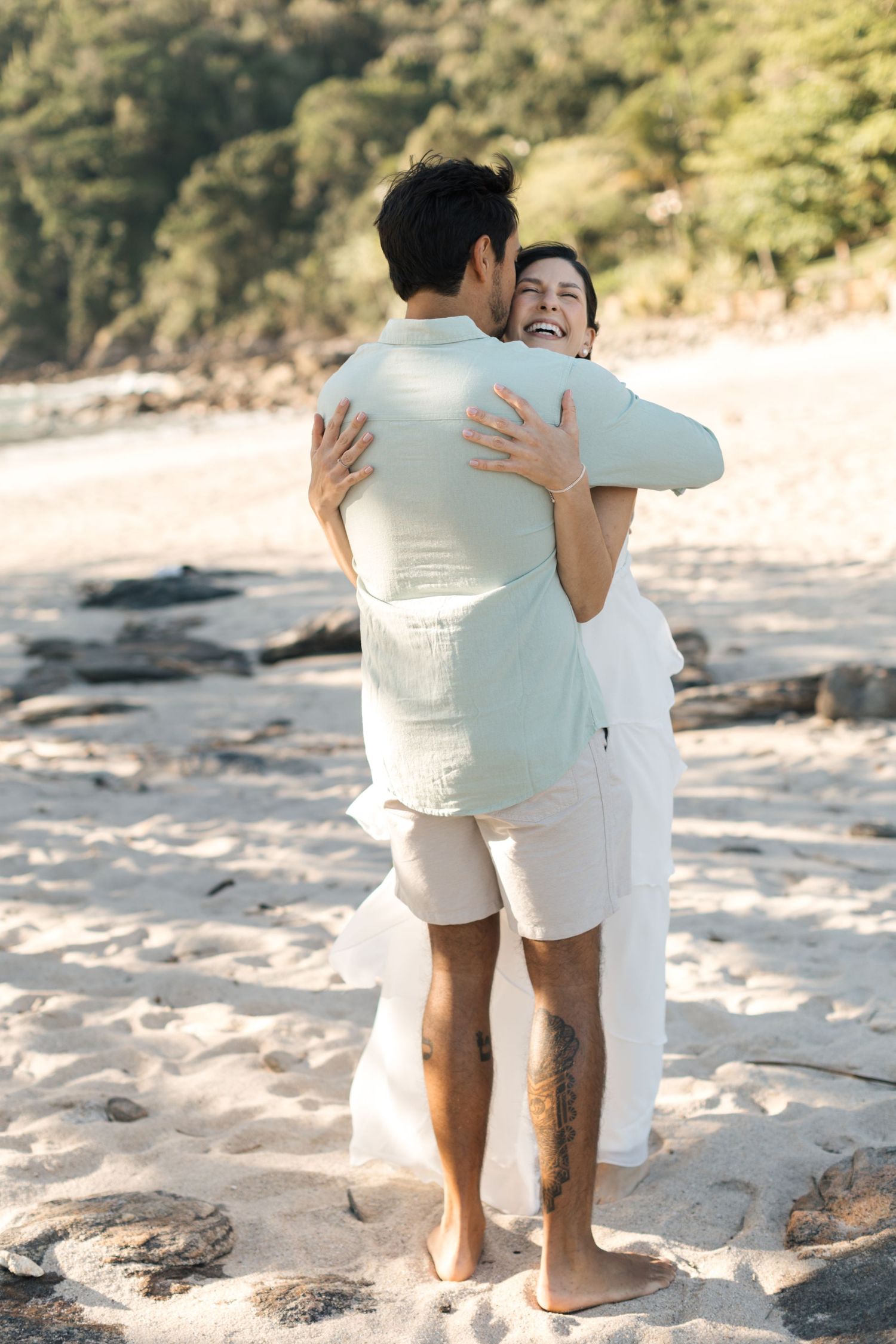 A couple in white clothing embraces while walking on a sandy beach at sunset.