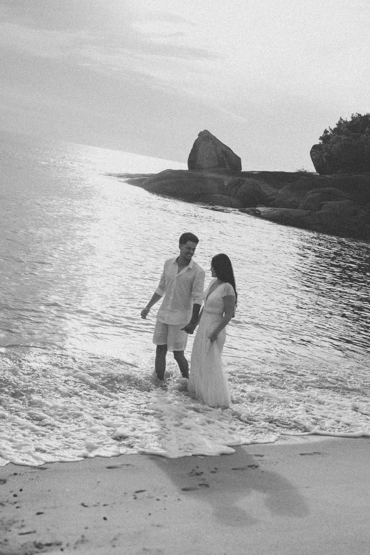 A couple stands in shallow ocean water at sunset near rocky coastal cliffs.