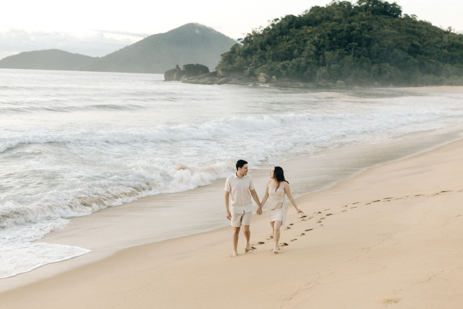 Couple walking hand in hand on a scenic beach with mountains and waves in the background.