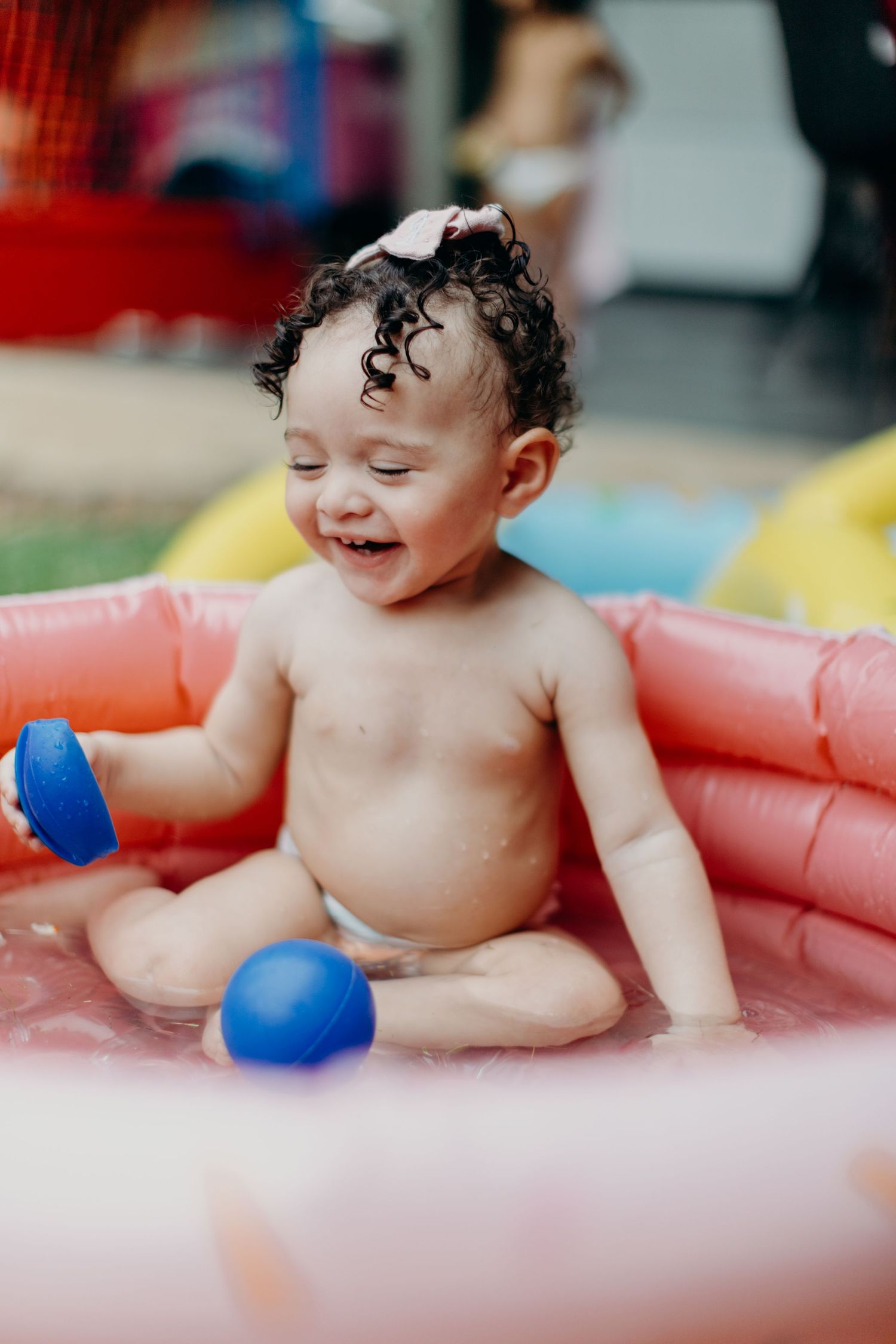 Happy baby playing with blue balls in pink inflatable pool.