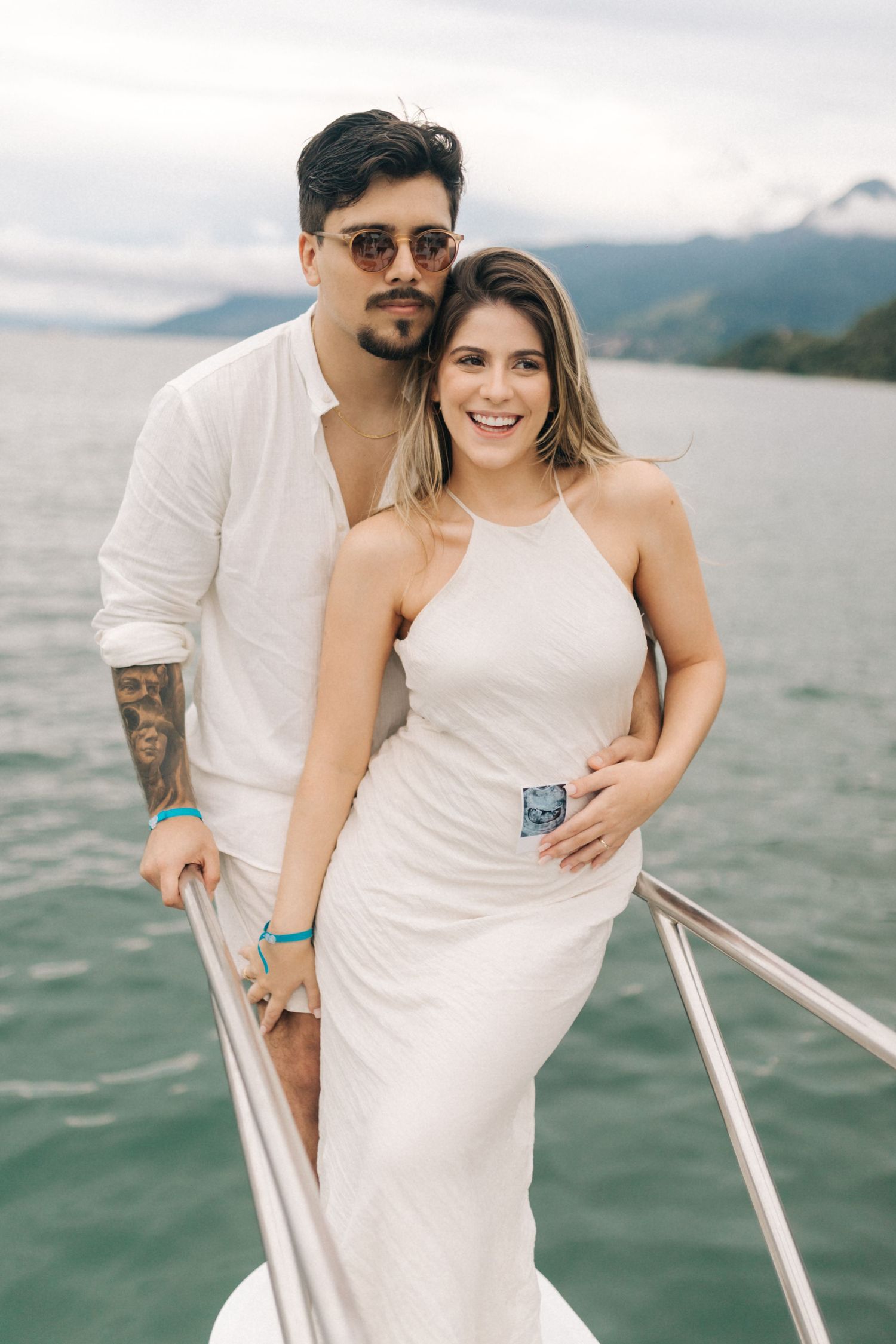 Couple in matching white outfits pose together on a boat deck with scenic mountain views.