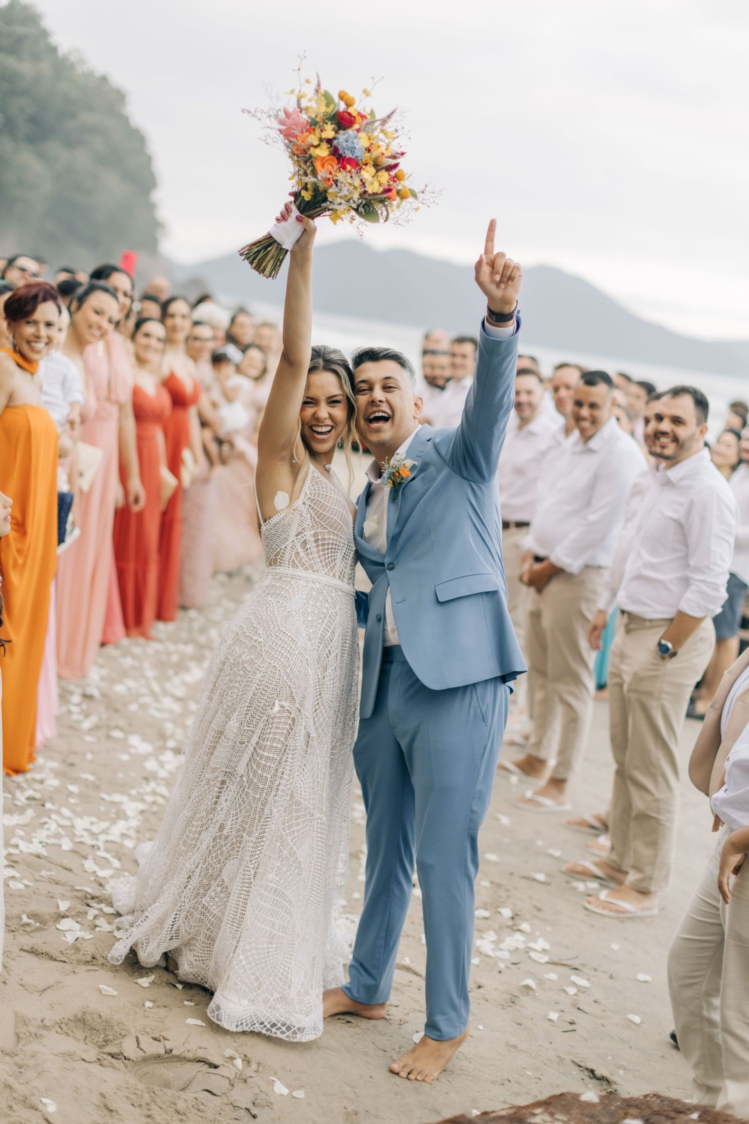 Newlyweds celebrate with raised arms as guests shower them with flower petals on beach.