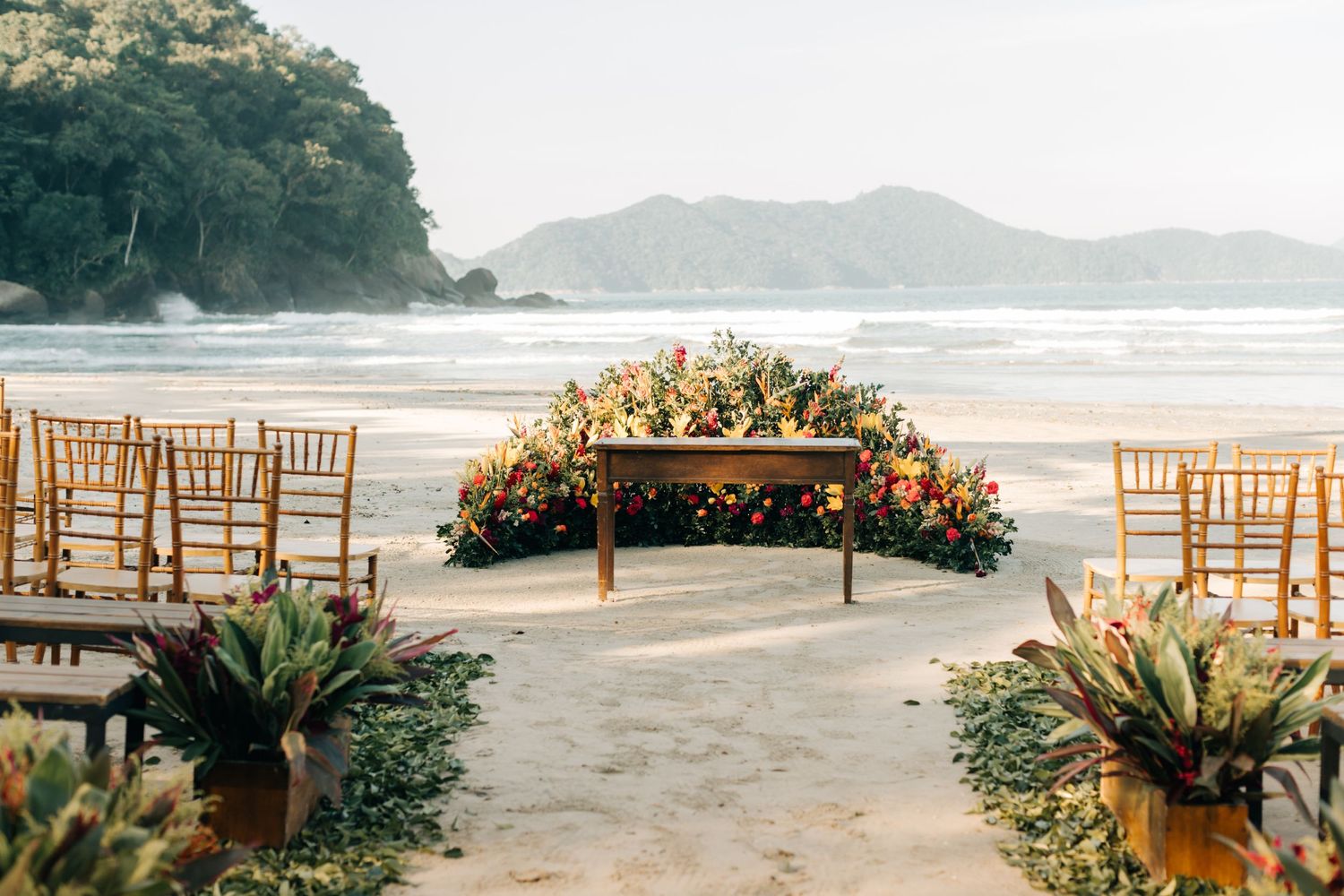 Beach wedding ceremony setup with wooden chairs and tropical floral arrangements.
