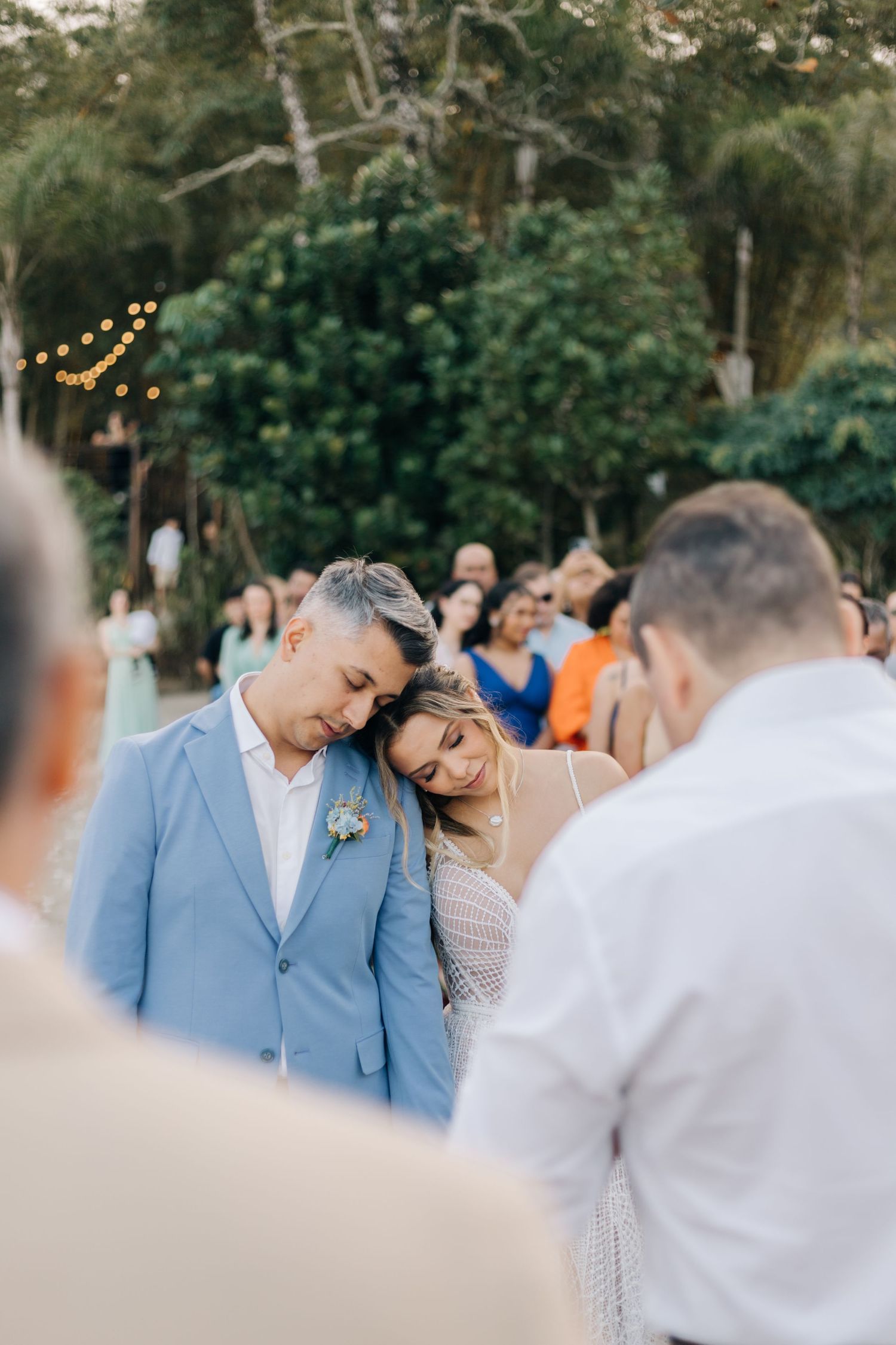 Intimate moment at an outdoor wedding ceremony as couple embraces under natural greenery and string lights.