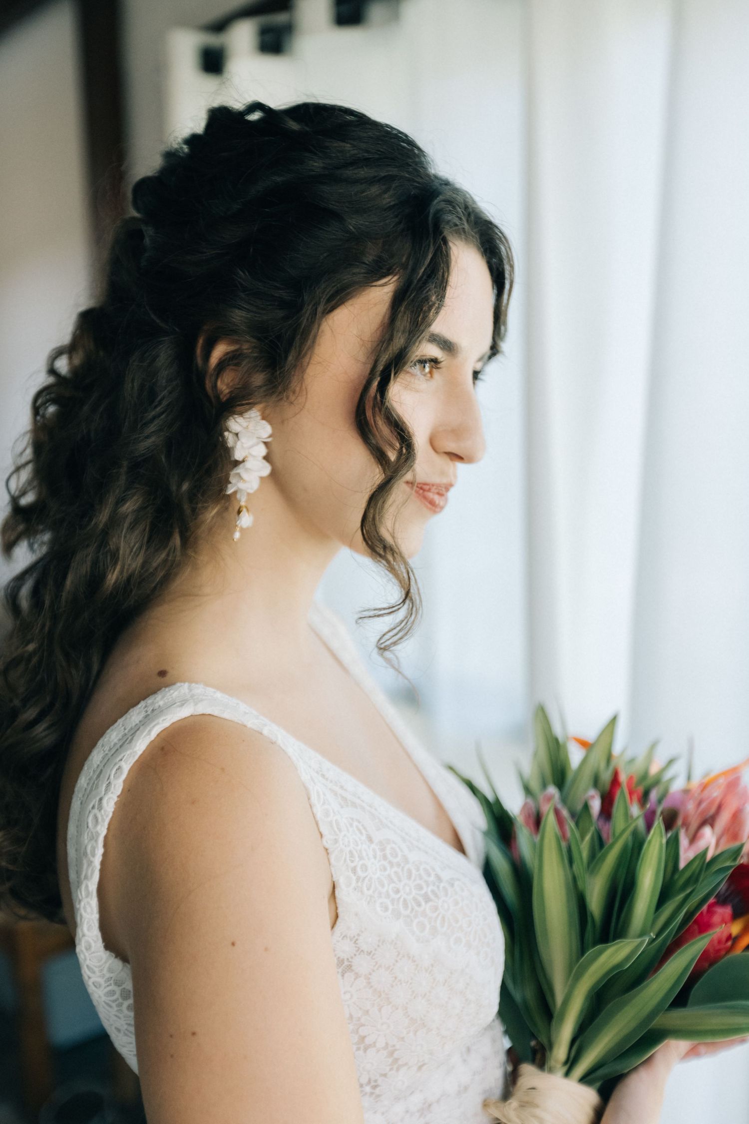 Bride in lace white gown holds tropical bouquet against white backdrop.