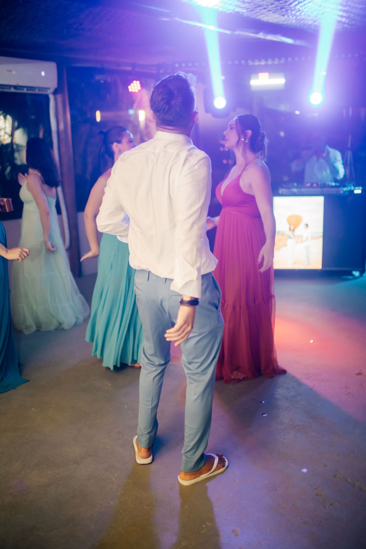 Wedding guests dance under purple lighting while wearing colorful formal dresses and suits.