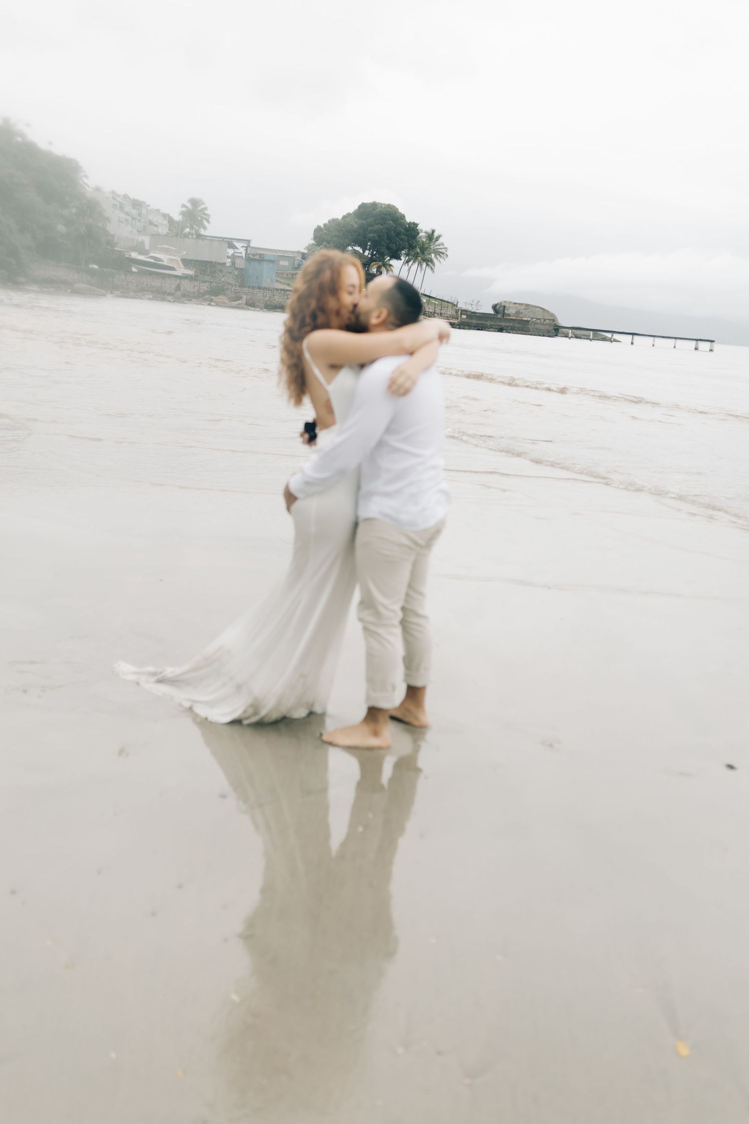 A couple in white attire shares an intimate embrace while standing barefoot on a foggy beach.