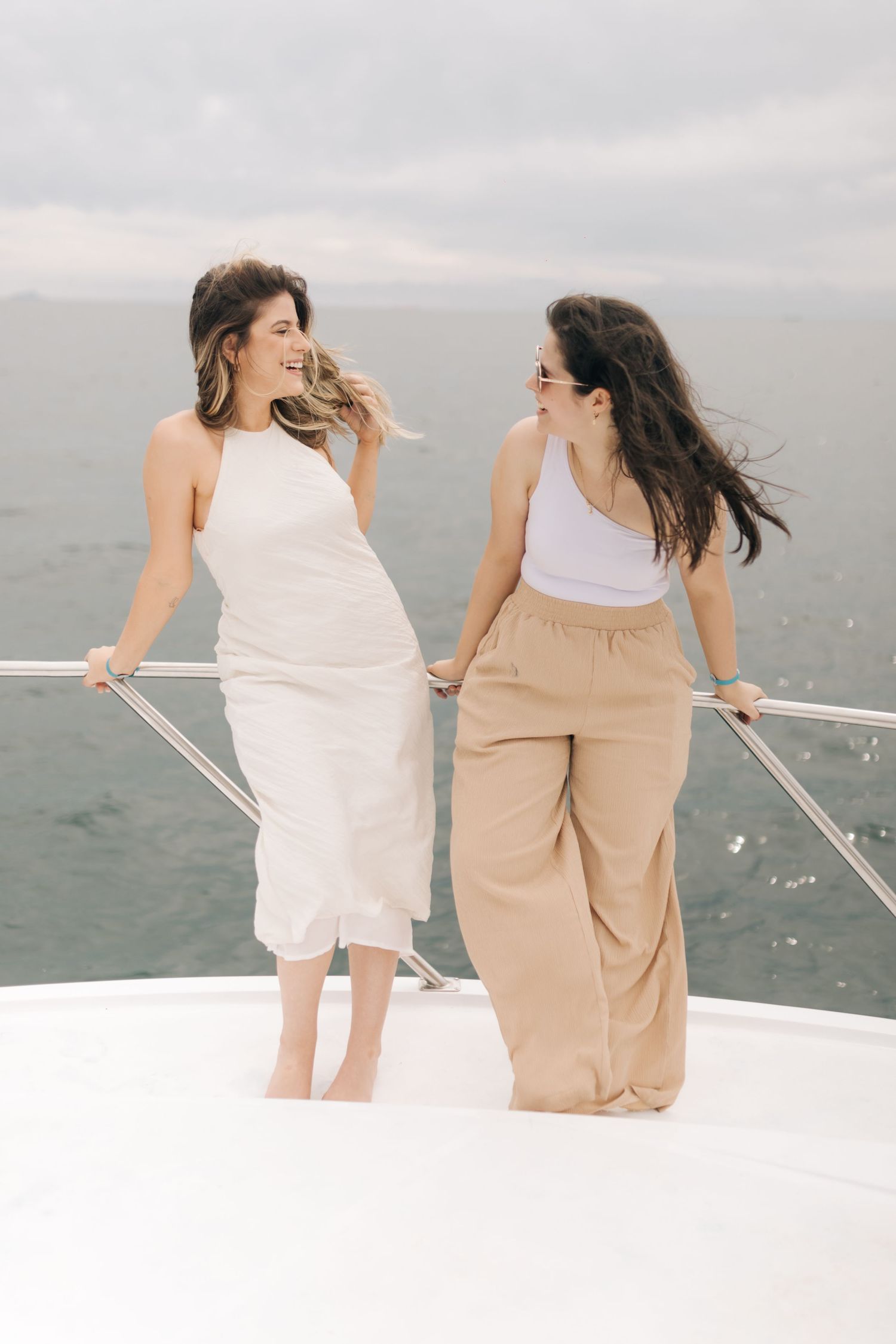 Two women in white and beige outfits enjoying the sea breeze on a yacht deck.