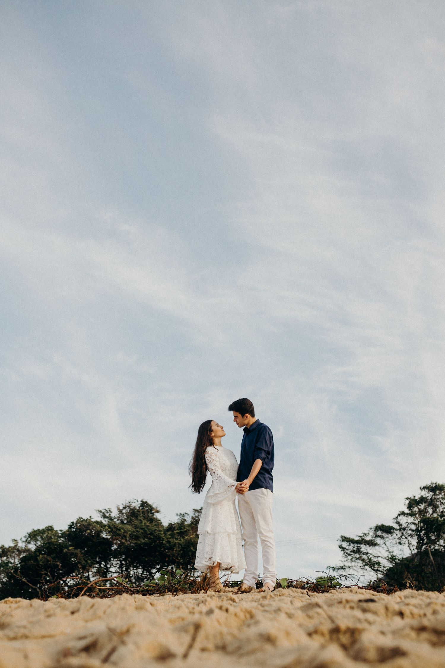 A couple shares an intimate moment while standing on sandy terrain under a bright blue sky.