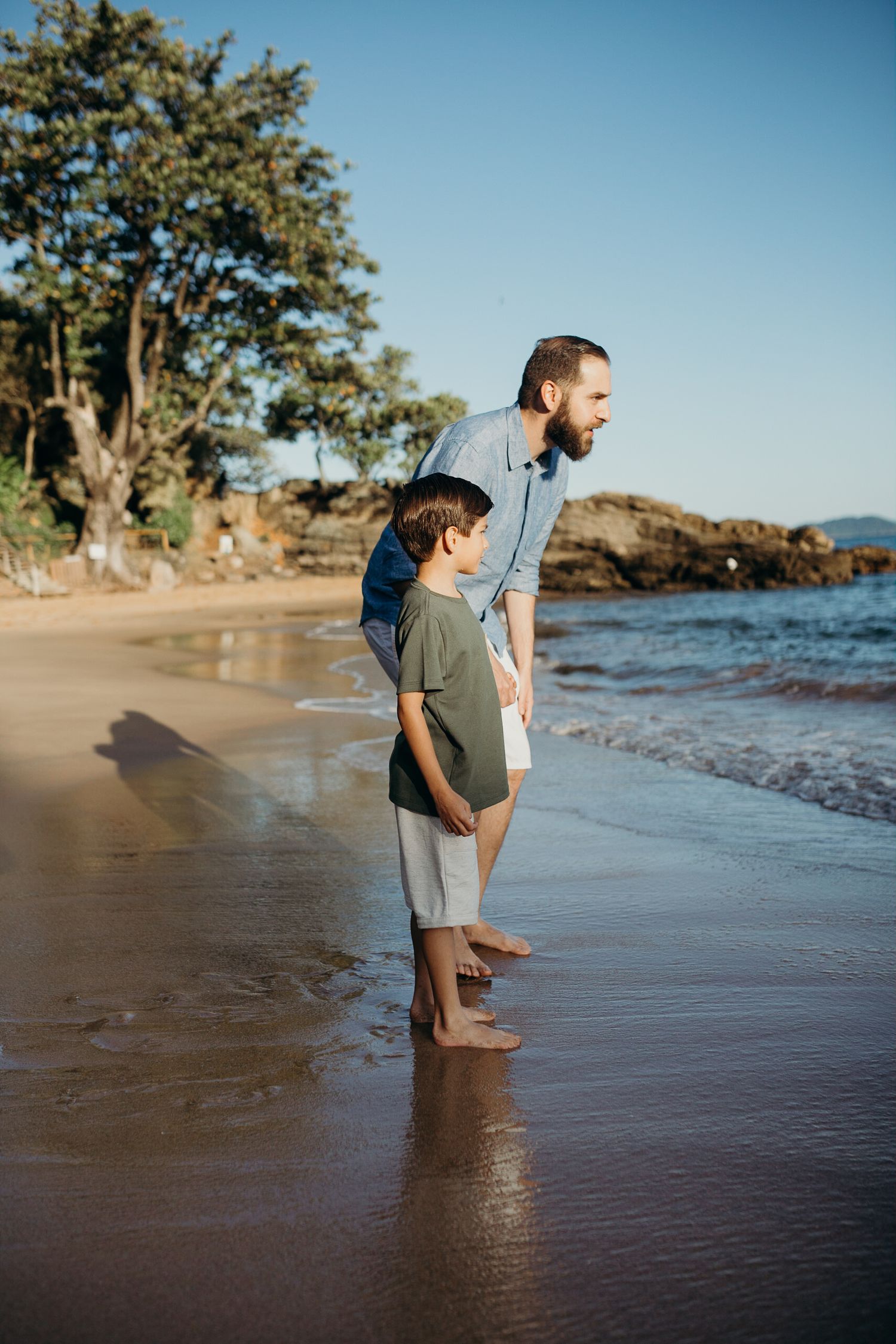 Two people standing at the edge of ocean waves on a scenic beach with trees.