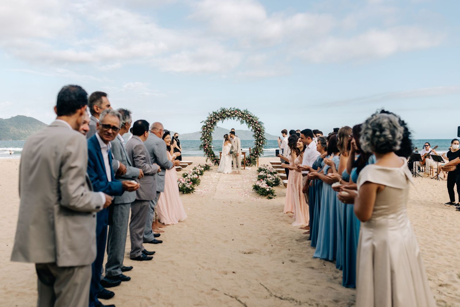 Wedding guests line the aisle on a beach as the ceremony begins under a floral arch.