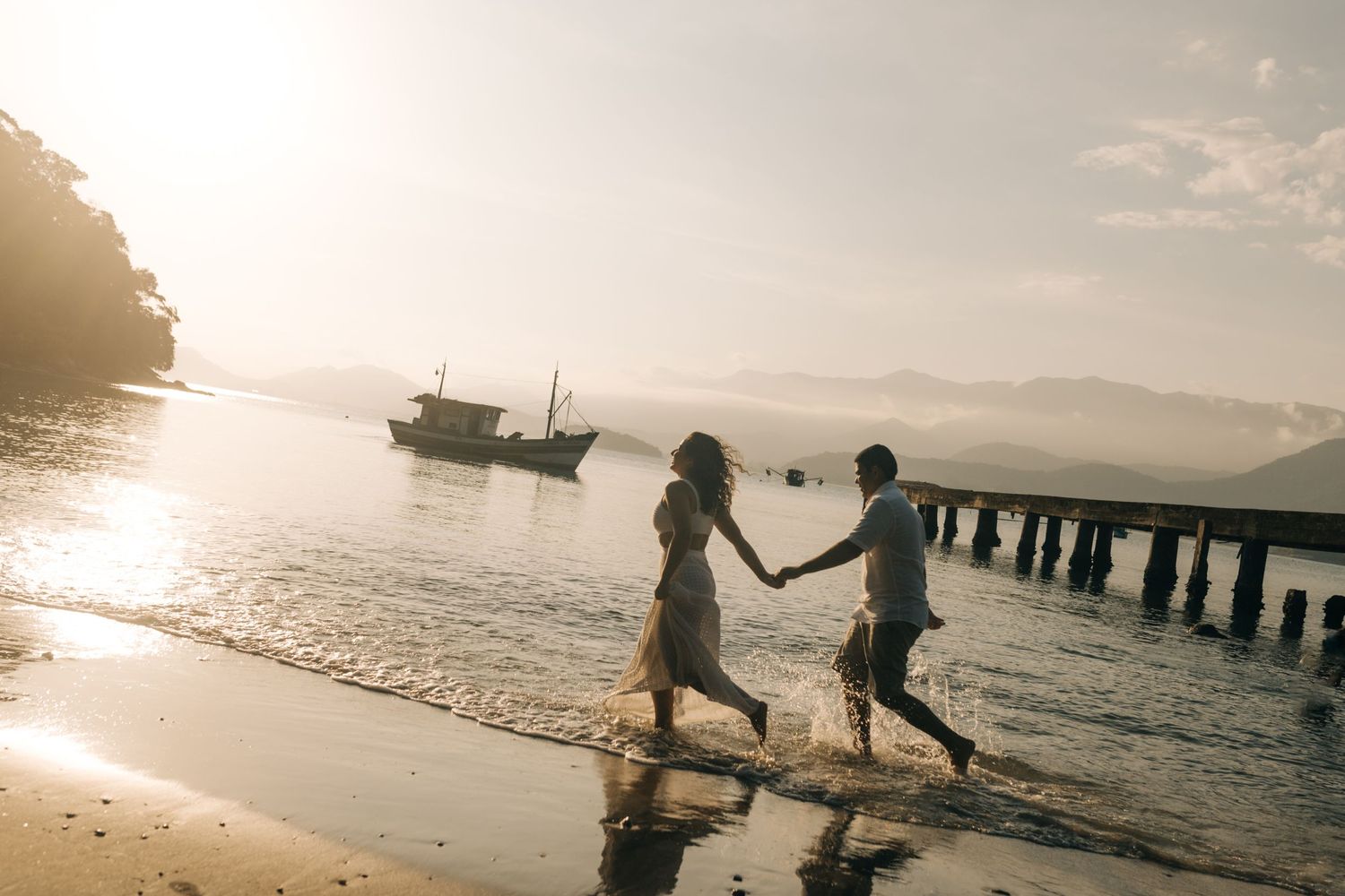 Silhouetted couple runs along beach shore at sunset with fishing boat in background.