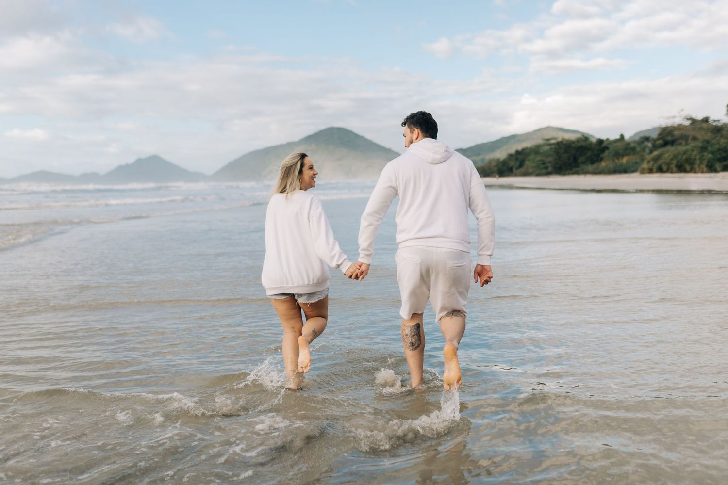 Couple walking hand in hand through shallow water with mountains in background.