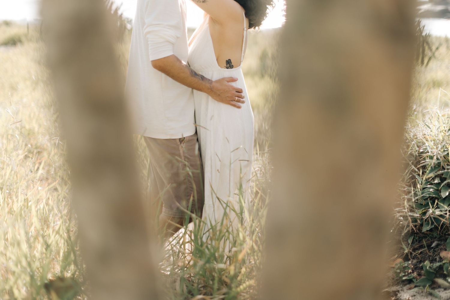 A couple dressed in white embraces while standing in tall golden grass during sunset.