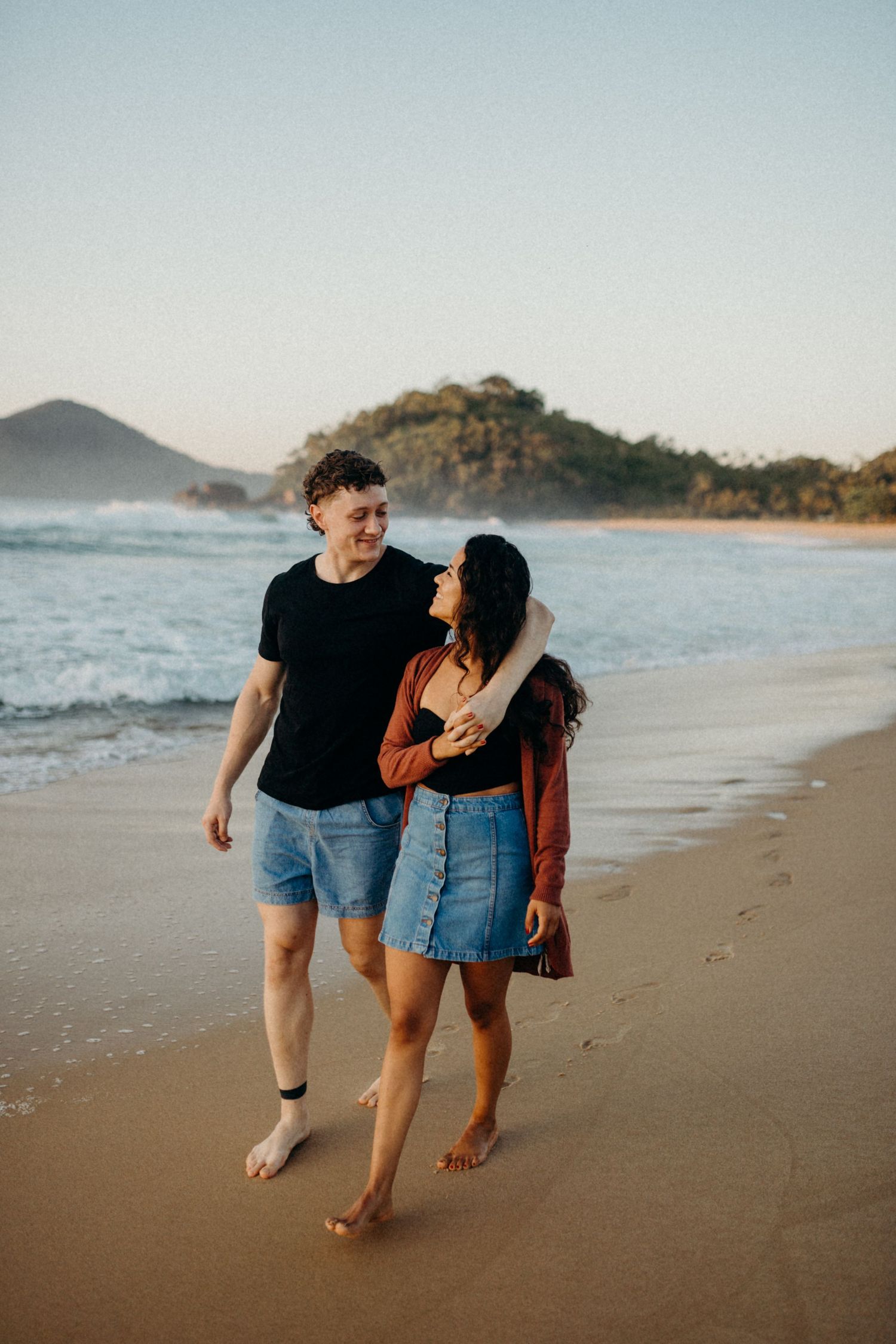 Casual couple walking barefoot along the beach at sunset with coastal mountains in view.